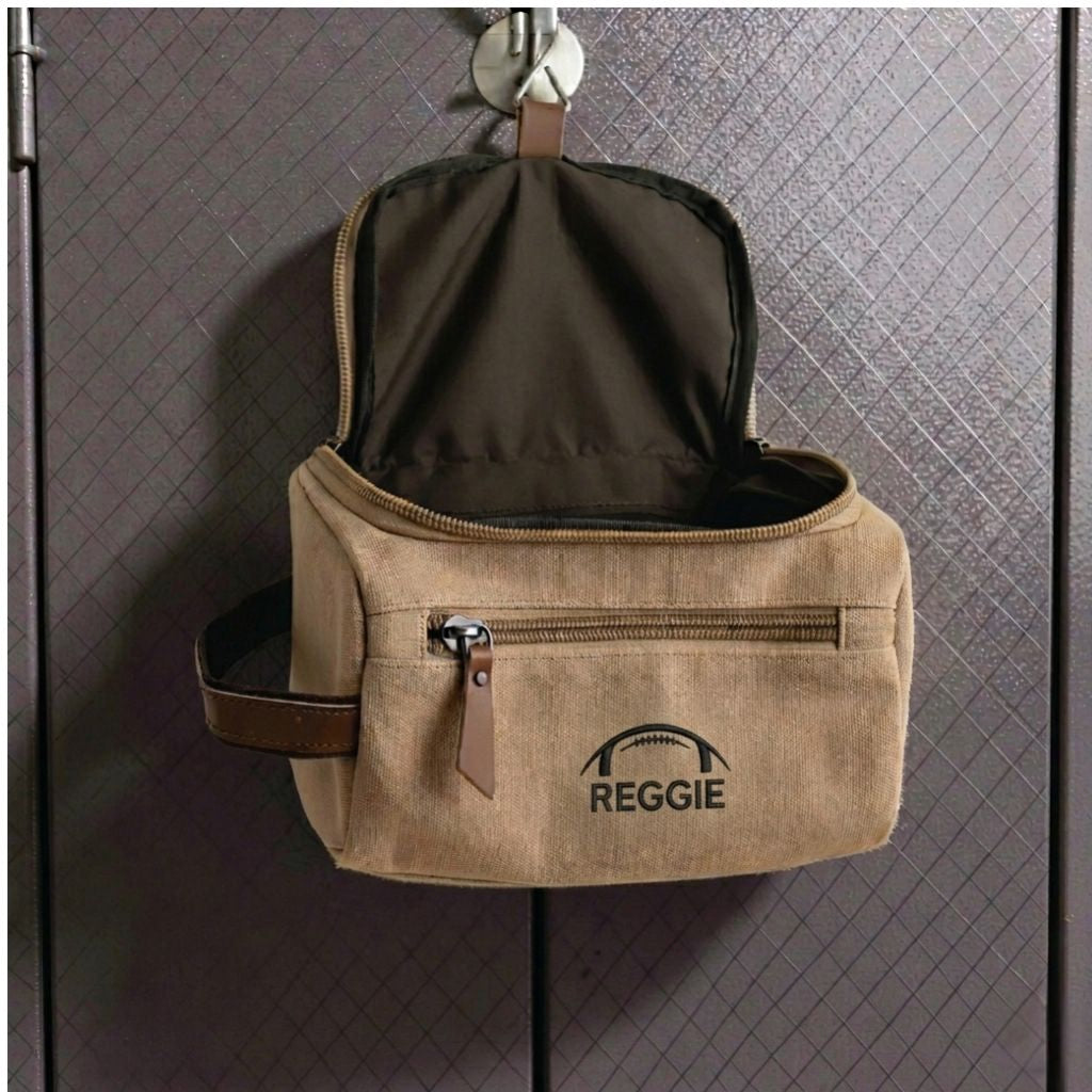 Tan canvas toiletry bag with leather handle, open to show black interior lining, personalized with sample name "REGGIE" and football embroidery on front