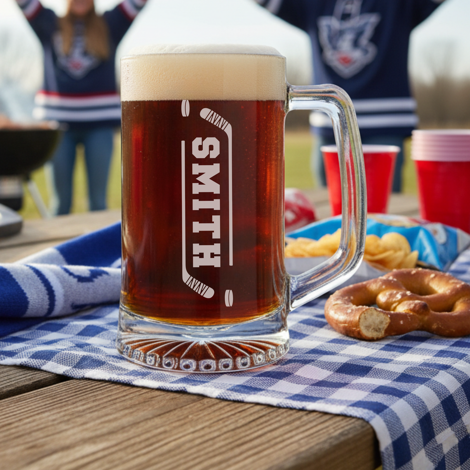 Personalized beer mug engraved with sample name "SMITH" between crossed hockey sticks, filled with dark beer and displayed at outdoor tailgate with snacks and sports fans in background