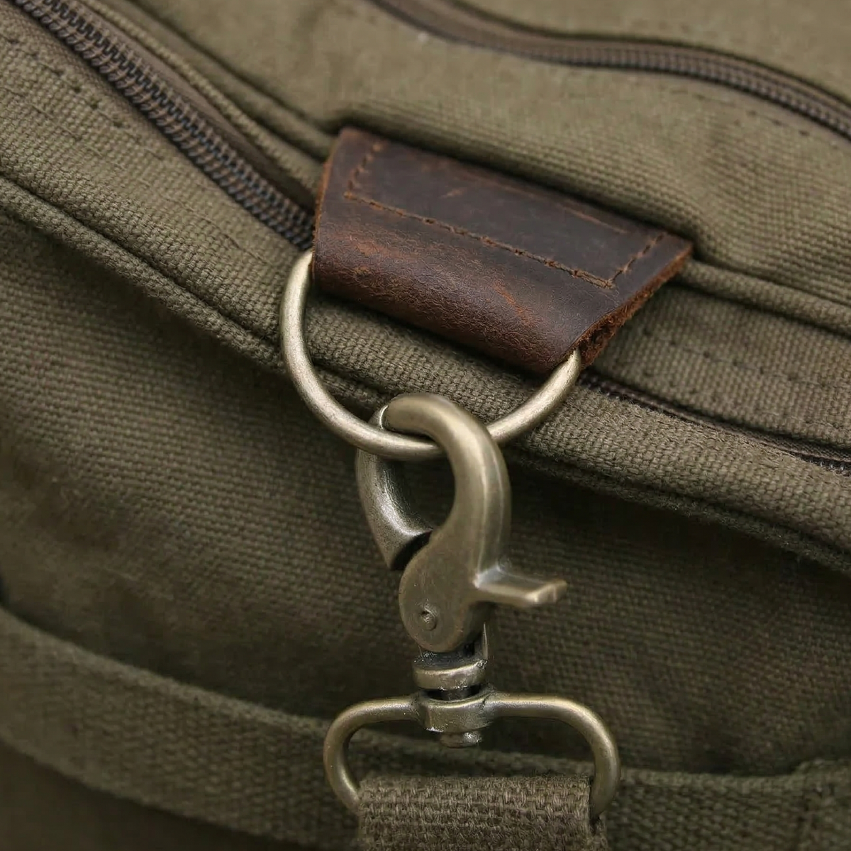 Canvas weekender bag in olive green with dark brown leather accent patch and metal snap hook detail attached to zippered pocket.