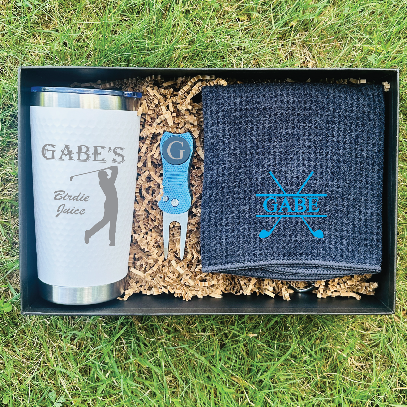Golf gift set displayed in black presentation box with straw fill, showing personalized tumbler engraved "GABE'S Birdie Juice," blue divot tool with magnetic marker, and embroidered navy towel with sample "GABE" monogram