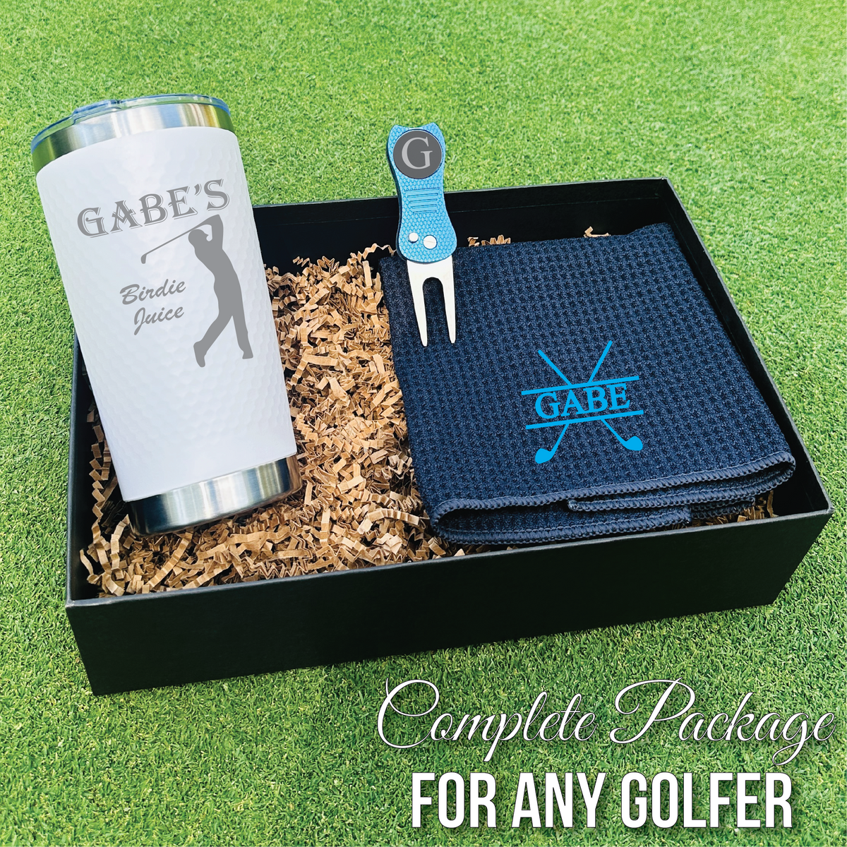 Golf gift set in black presentation box containing personalized white tumbler engraved "Gabe's Birdie Juice," navy towel with sample monogram "GABE," blue divot tool, and ball marker on green turf background