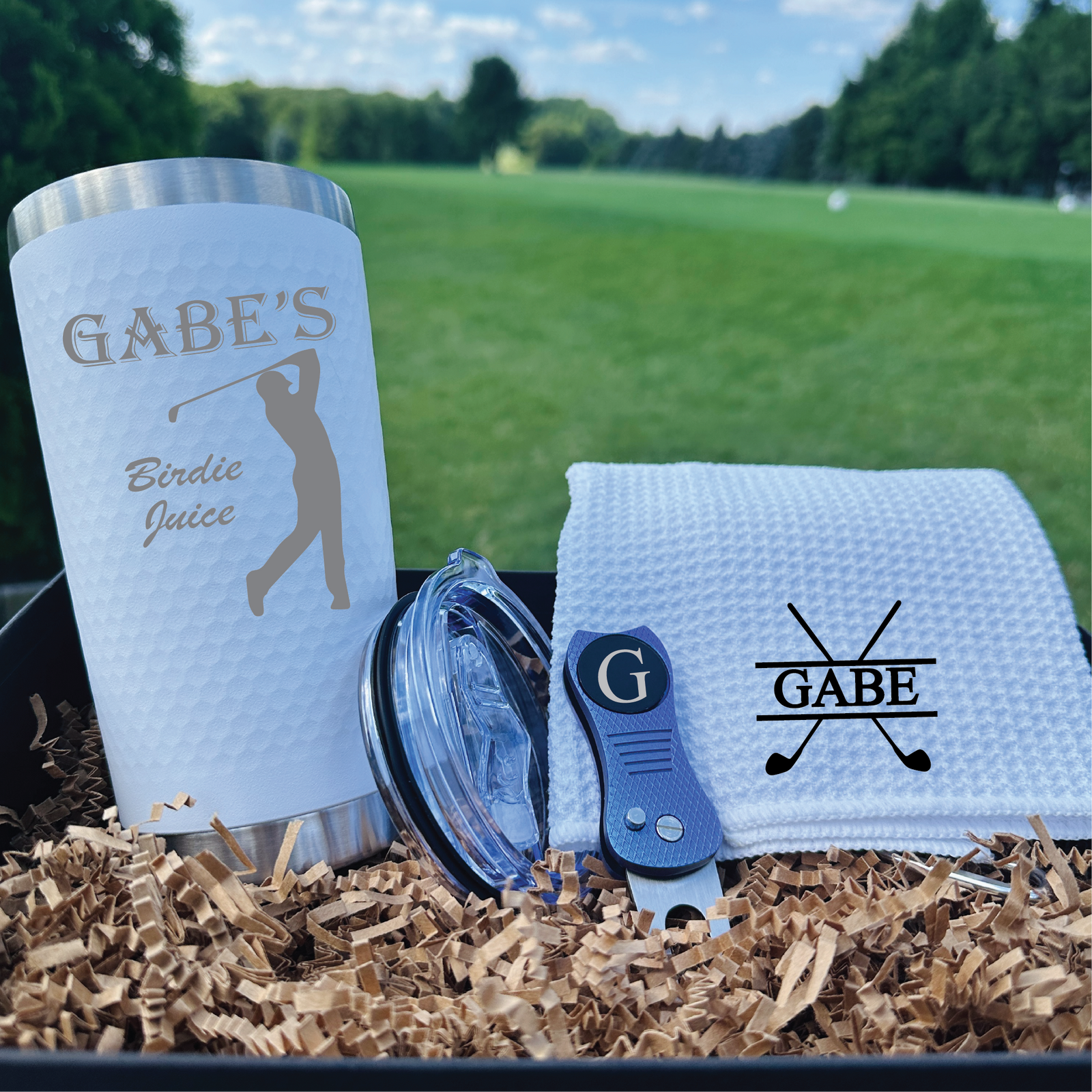 Personalized golf set displayed on grass showing engraved tumbler with sample name "GABE'S" and "Birdie Juice" text, embroidered towel with crossed-clubs motif, divot tool, and ball marker on packing material with golf course background