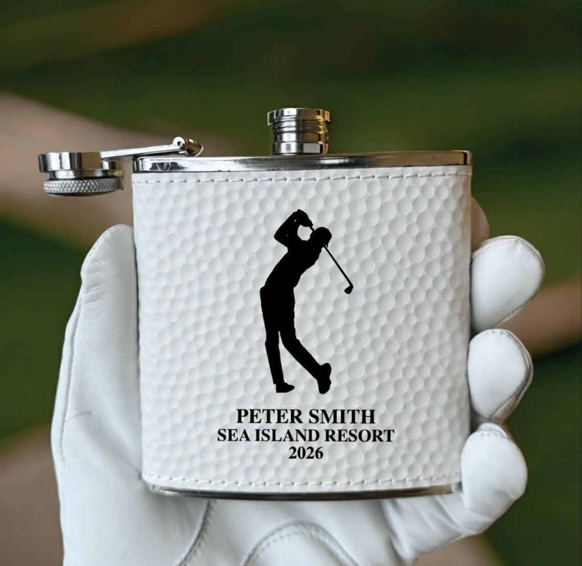 White golf outing gift flask with golf-themed design held in a white glove against a blurred green background