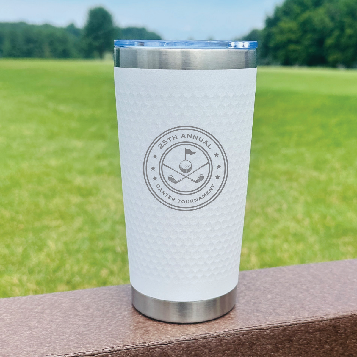 White insulated tumbler with stainless steel base and blue lid, engraved sample showing "26th Annual Golf Tournament" circular logo with golf flag and clubs