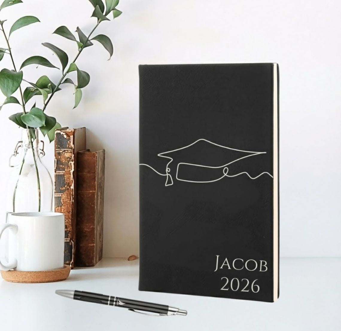 Black notebook with a graduation cap design and 'JACOB 2026' text, placed on a white surface with a plant and books in the background.