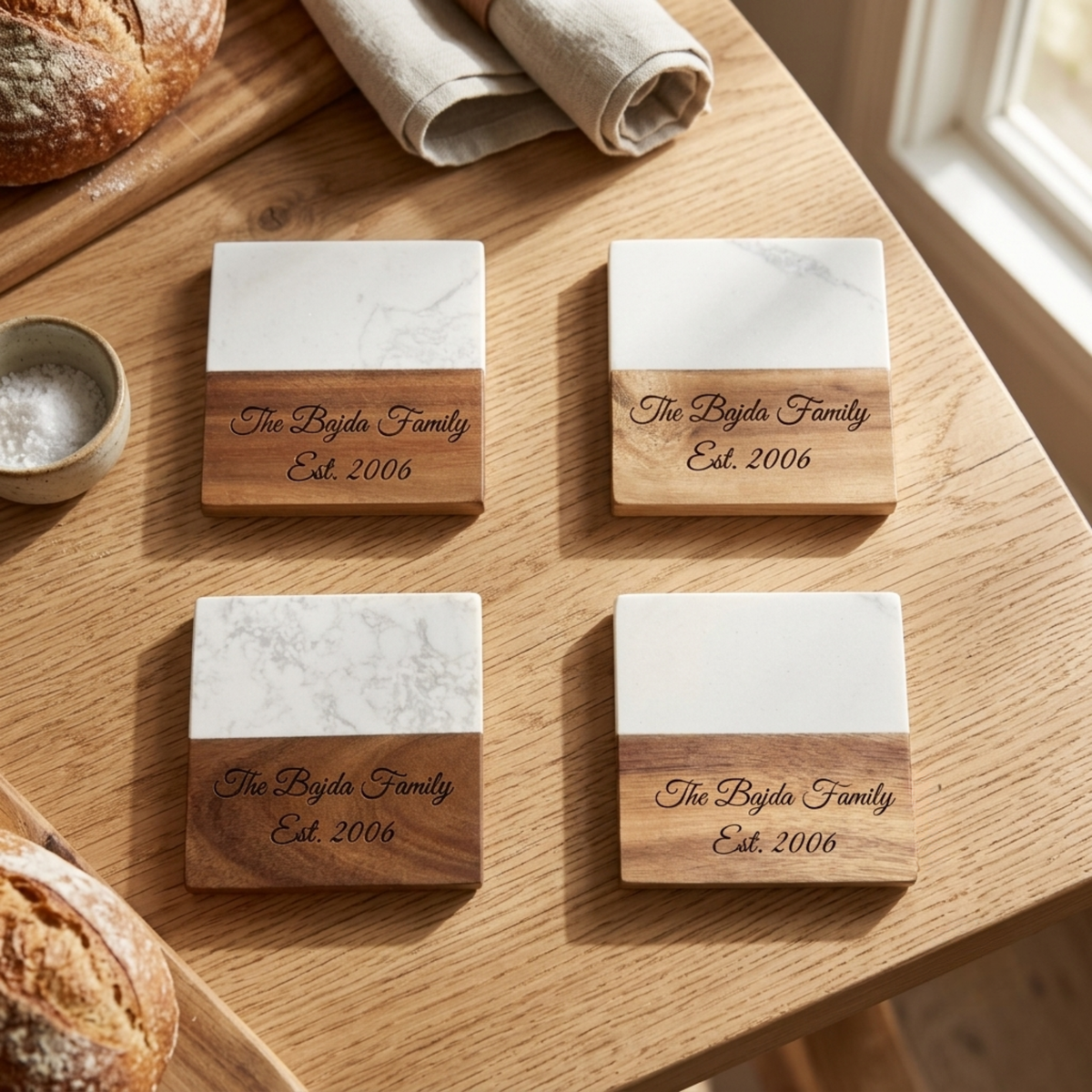 Four square coasters with white marble tops and engraved acacia wood bases showing a sample family name and established year, arranged on a wood surface.