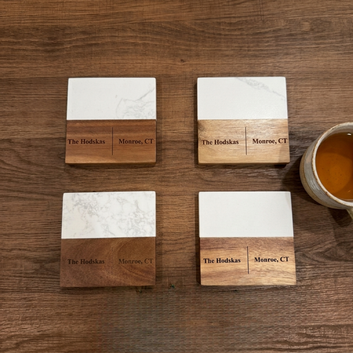 Four personalized coasters with white marble tops and acacia bases engraved with a sample family name and their city and state, arranged beside a drink.