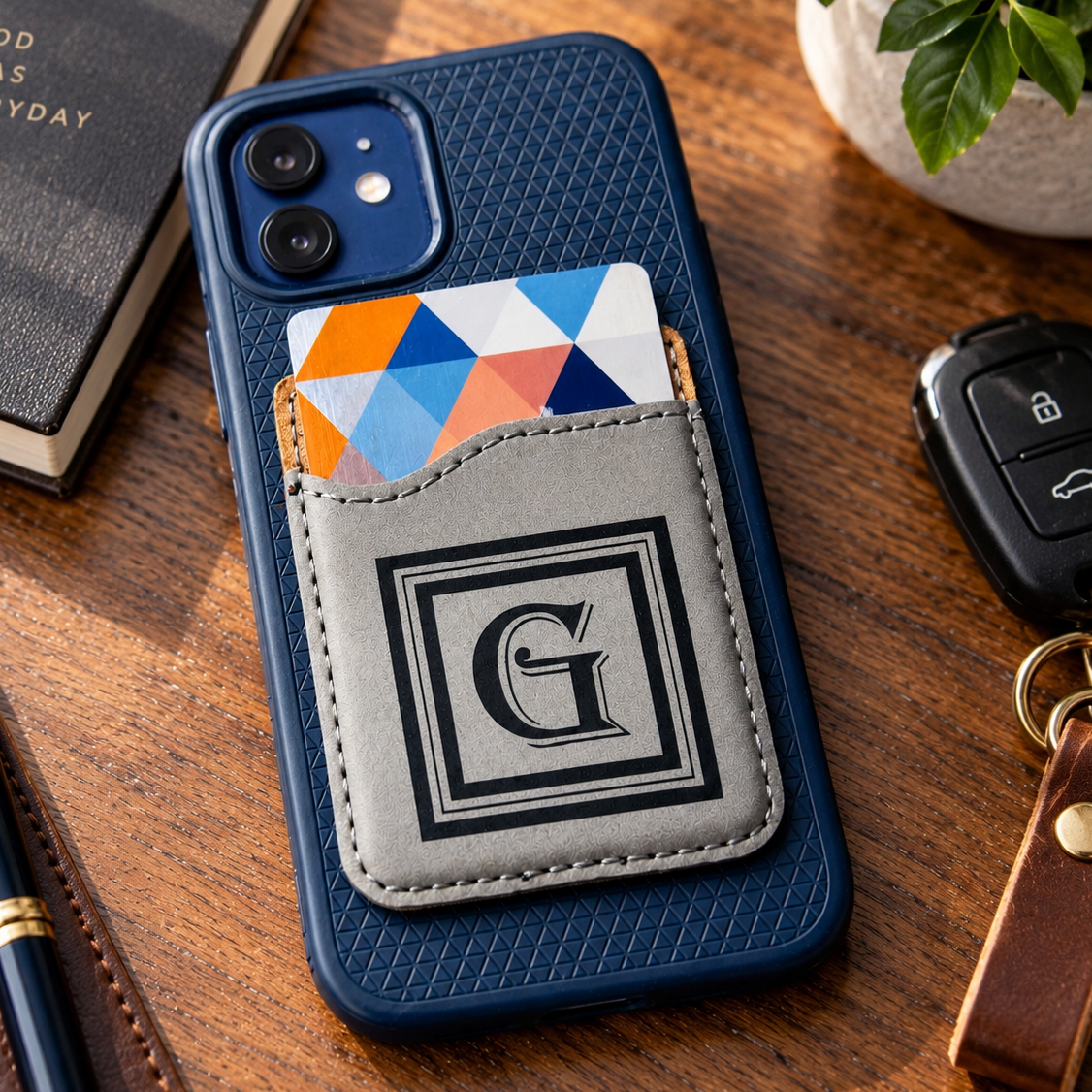 Monogram Phone Card Holder in gray leatherette engraved with a square G monogram, stuck to the back of a navy phone case near keys and a journal