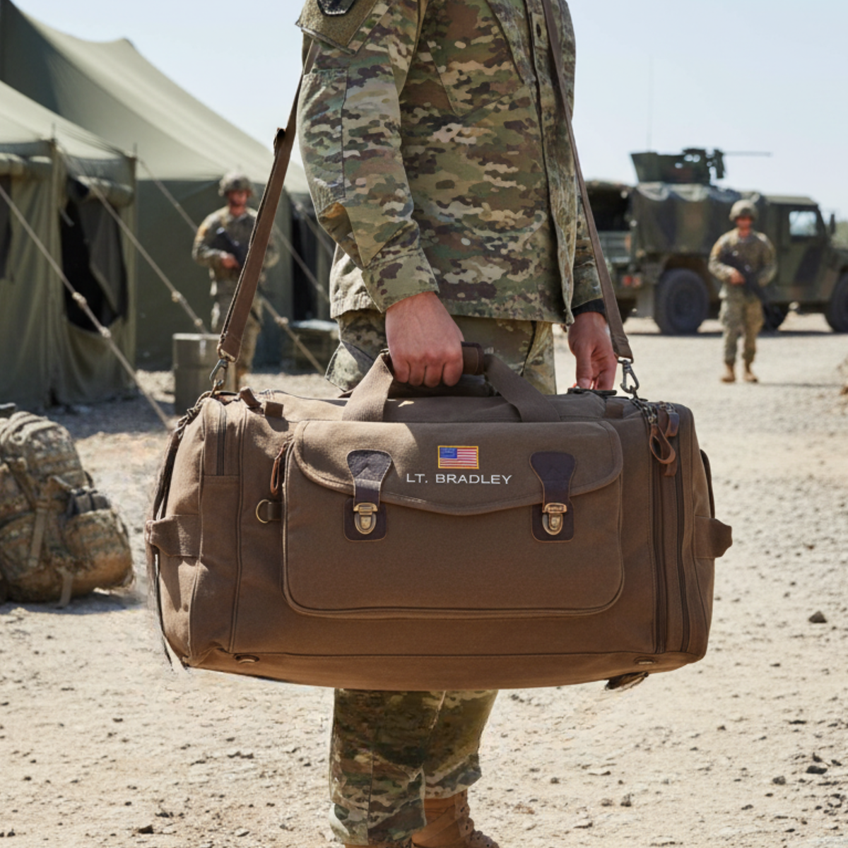 Brown canvas duffle bag personalized with sample engraving "LT. BRADLEY" and US flag patch, carried by person in camouflage uniform at military camp.