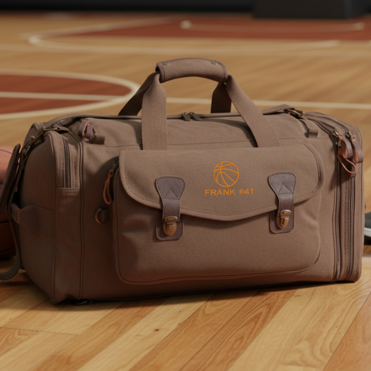 Tan canvas duffle bag with orange basketball logo and "FRANK 41" text, displayed on wooden basketball court floor with leather handles and brass hardware visible