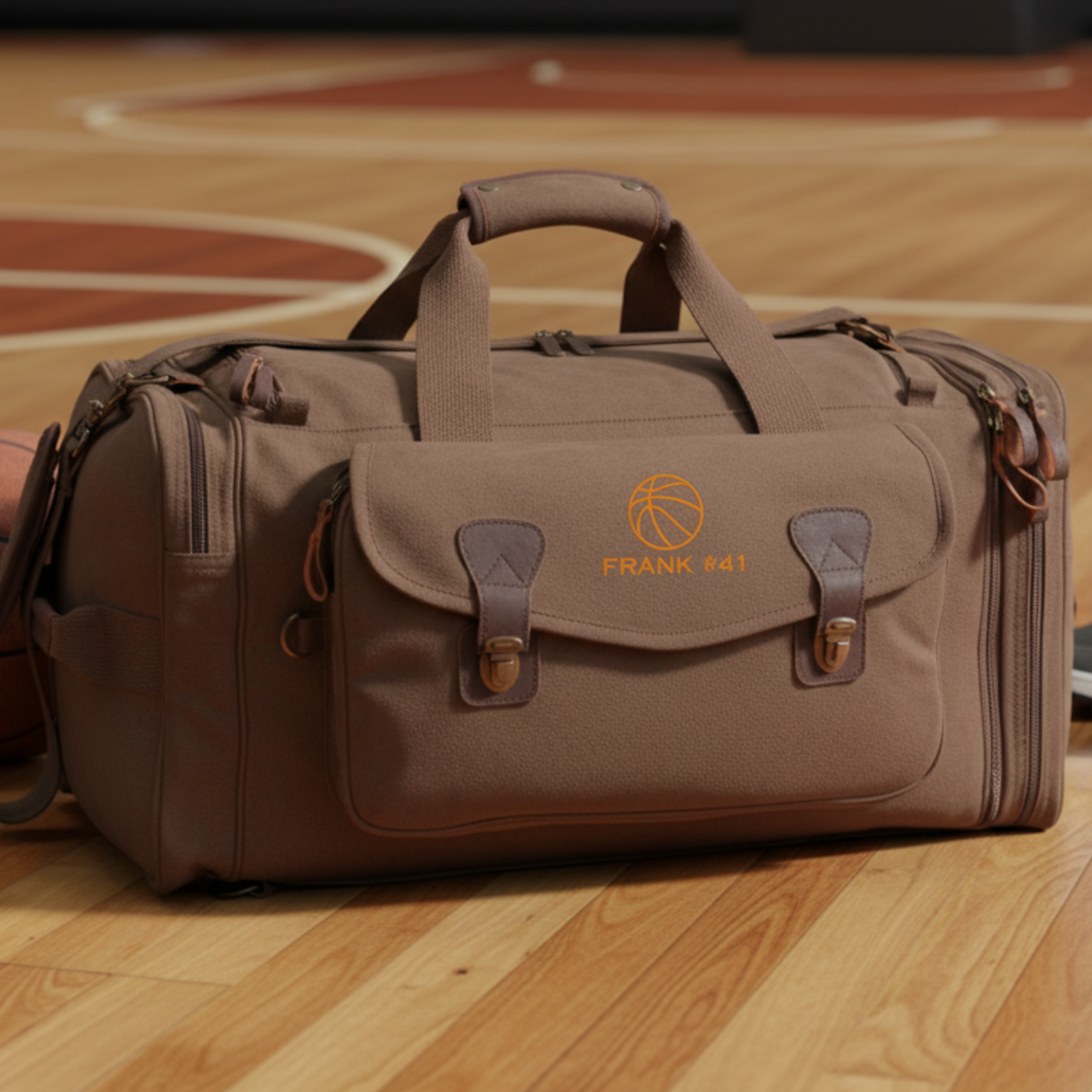 Tan canvas duffle bag with orange basketball logo and "FRANK 41" text, displayed on wooden basketball court floor with leather handles and brass hardware visible