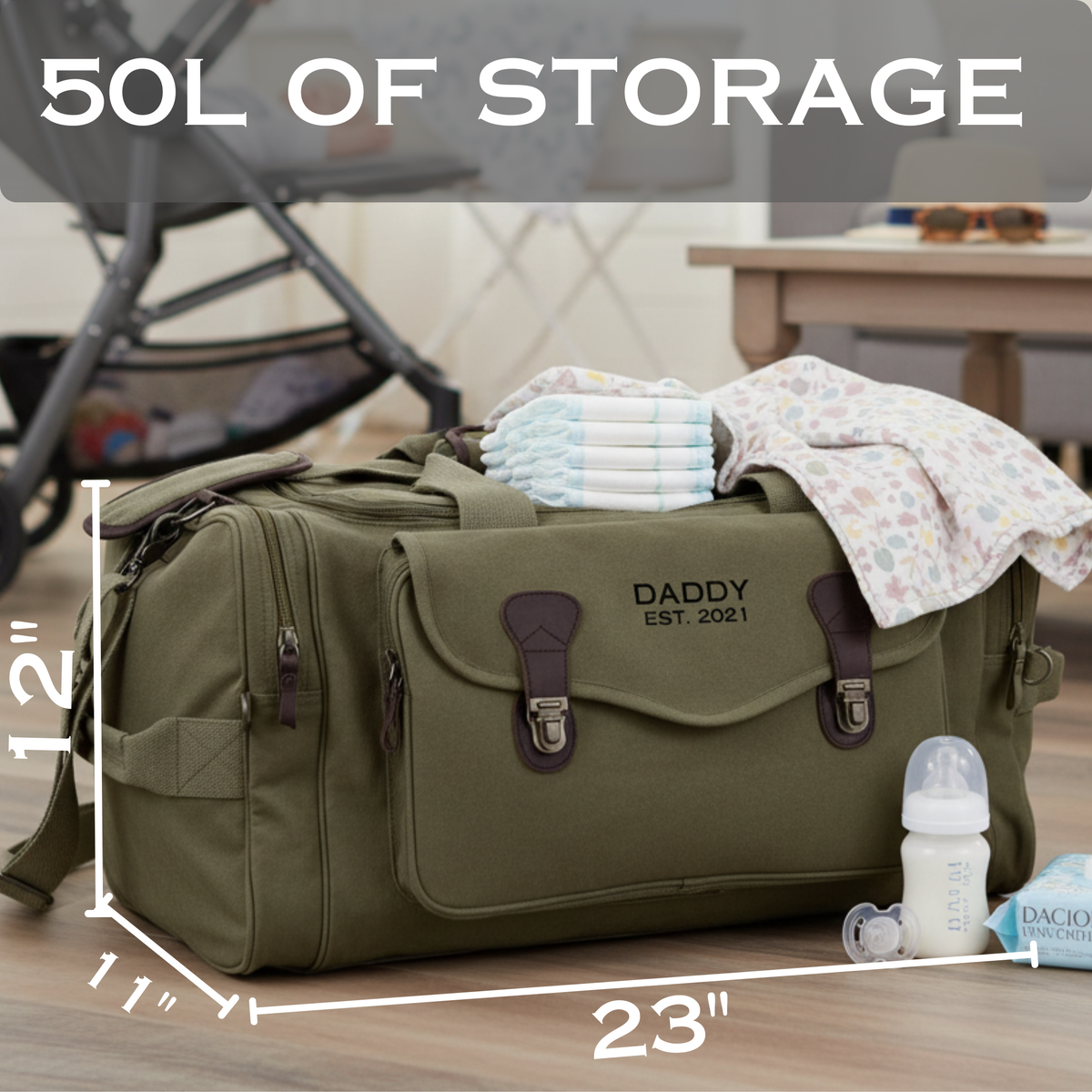 Olive green canvas duffle with embroidered sample text "DADDY EST. 2021" and brown leather trim, styled with diapers, blanket, and baby bottle on top