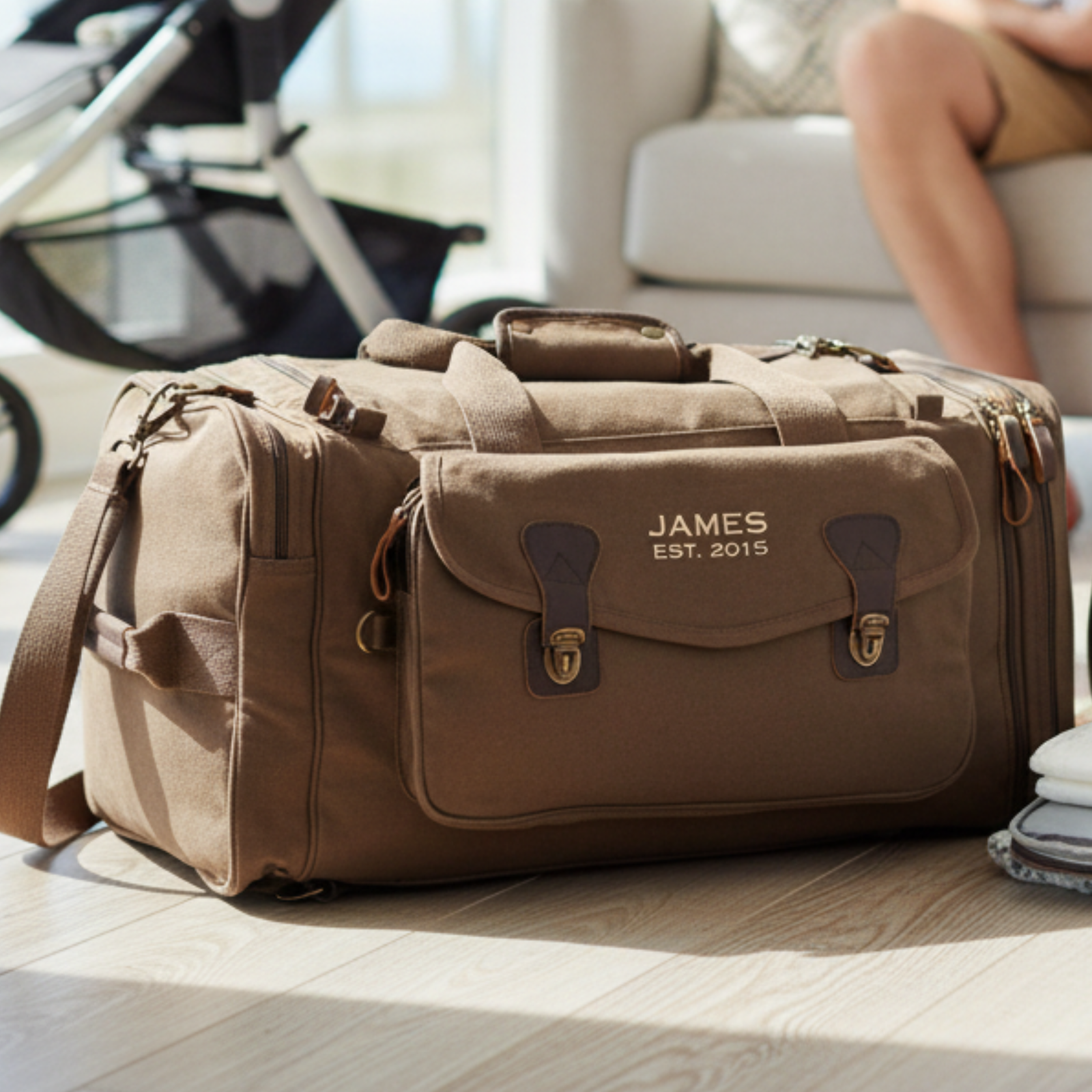 Tan canvas duffle bag with leather trim and brass hardware, personalized with sample engraving reading "JAMES EST. 2015" on front flap, displayed on wooden floor with shoulder strap visible