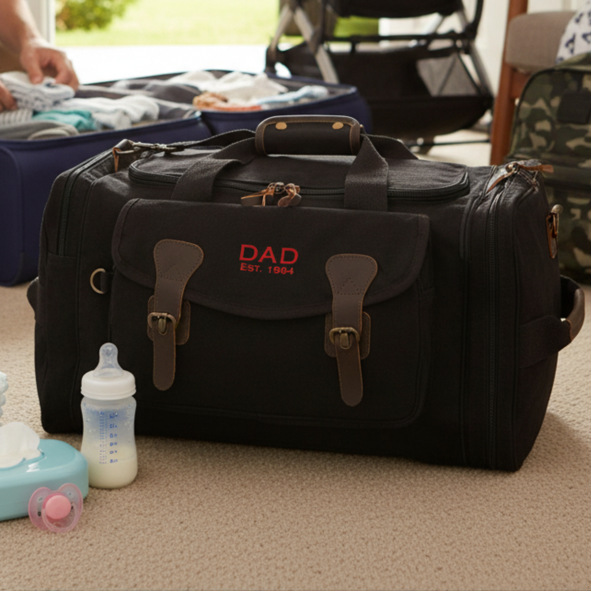 Black canvas duffle with brown leather handles, embroidered with sample text "DAD EST. 1904" in red, displayed on carpet surrounded by baby items