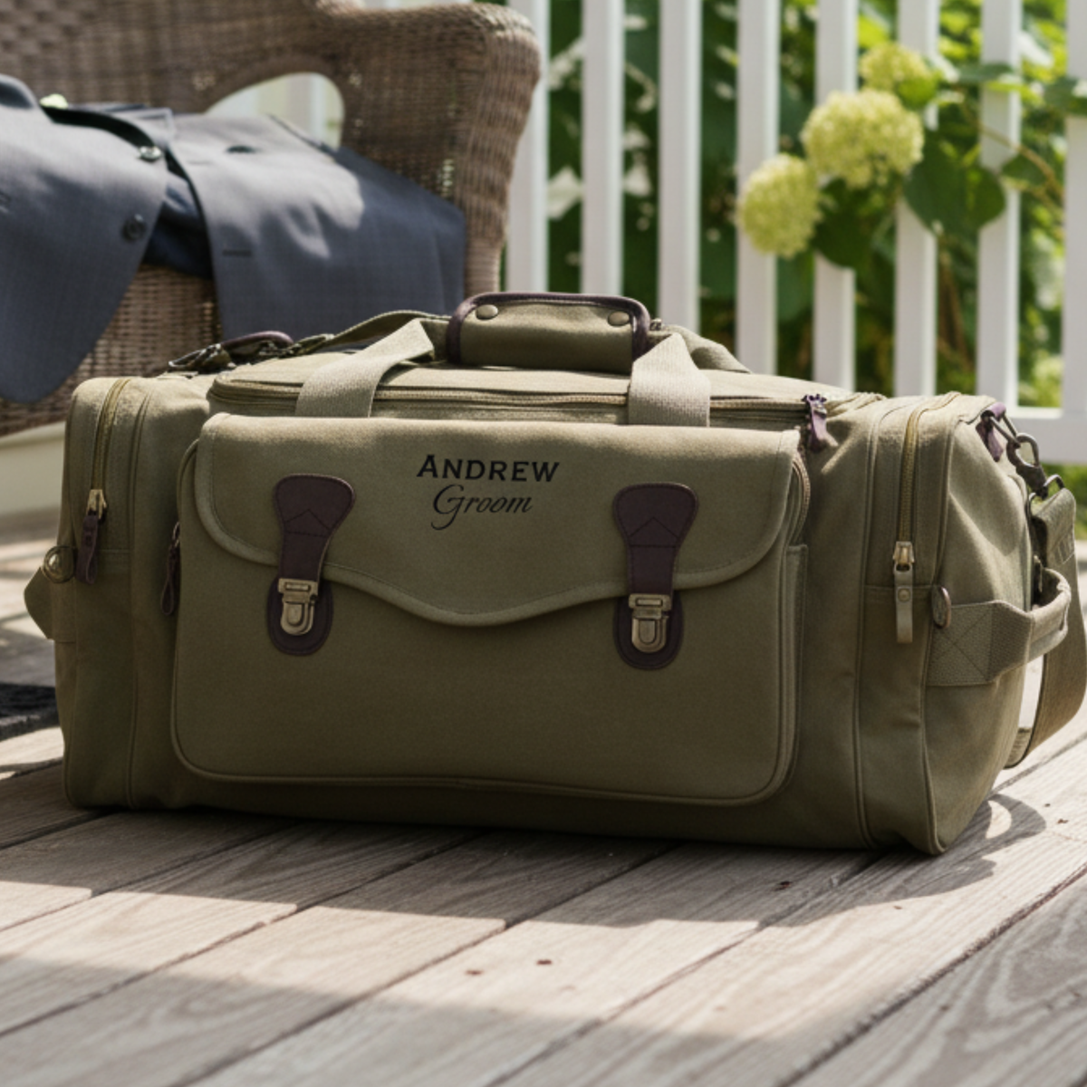 Tan canvas duffel bag personalized with sample text "ANDREW Groom" on front, burgundy leather straps and handles, displayed on wooden deck with garden background.