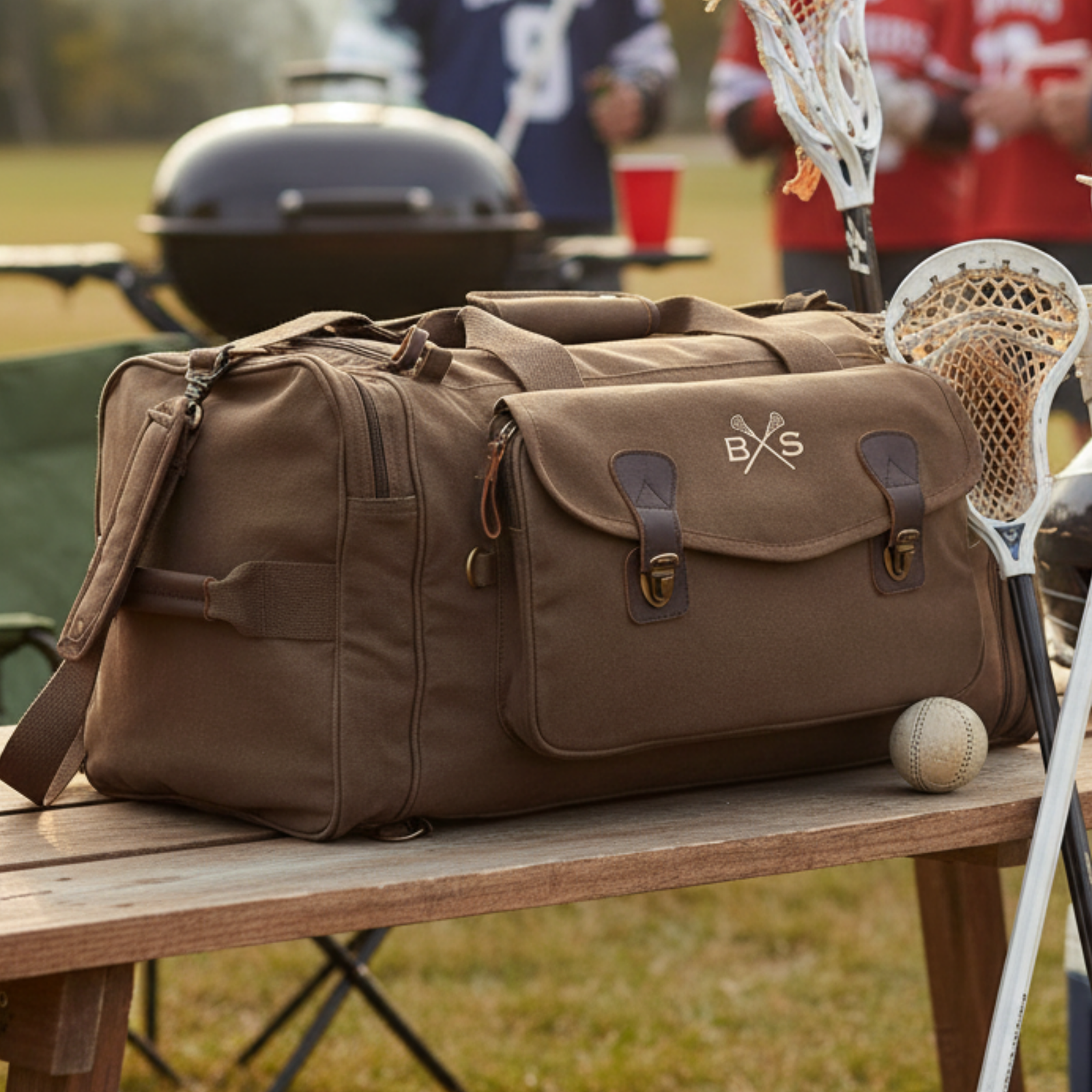 Brown canvas lacrosse duffle bag with leather accents and crossed-sticks logo, displayed on wooden table at outdoor sports field with equipment nearby
