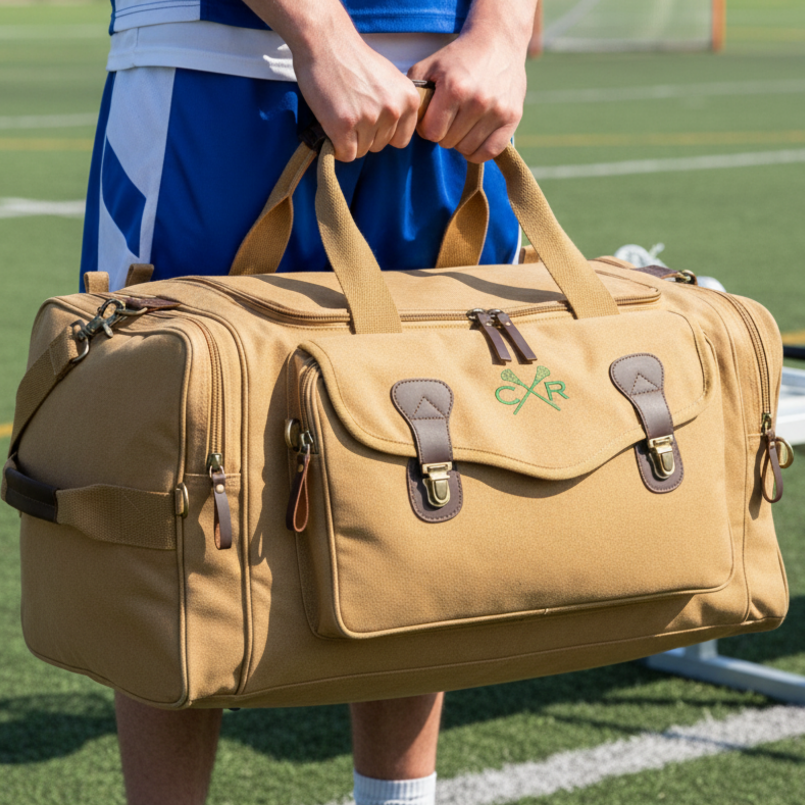 Tan canvas duffle bag with brown leather accents and crossed lacrosse sticks embroidered in green, held by person in blue athletic wear on sports field