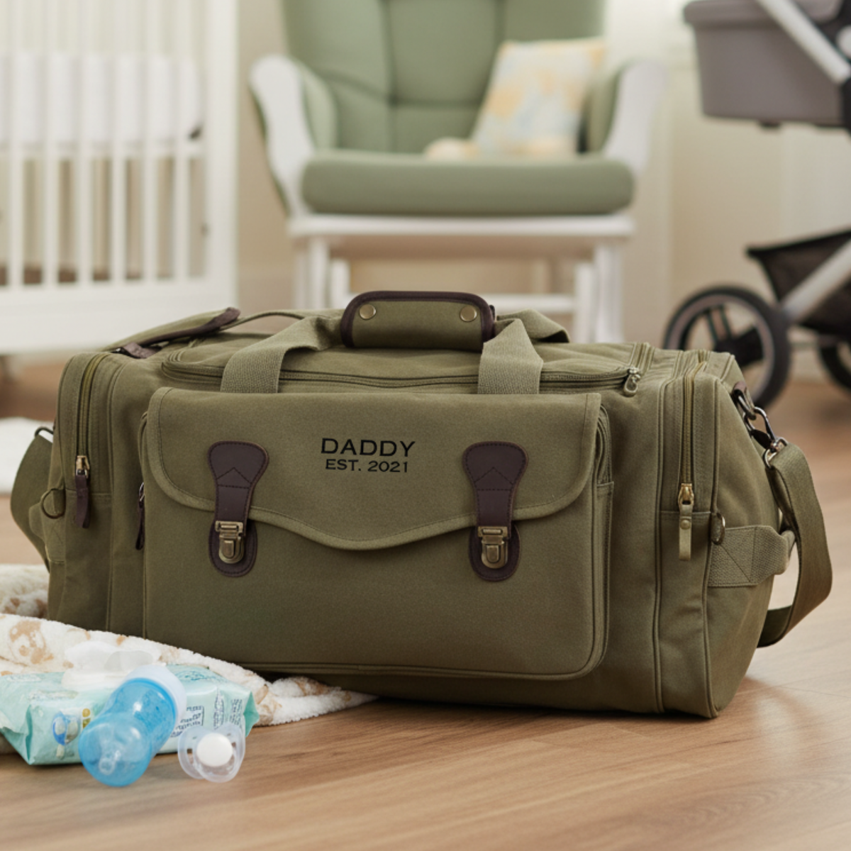 Olive green canvas duffle with leather trim, personalized with sample text "DADDY EST. 2021" embroidered on front flap, positioned on wooden floor with nursery items nearby