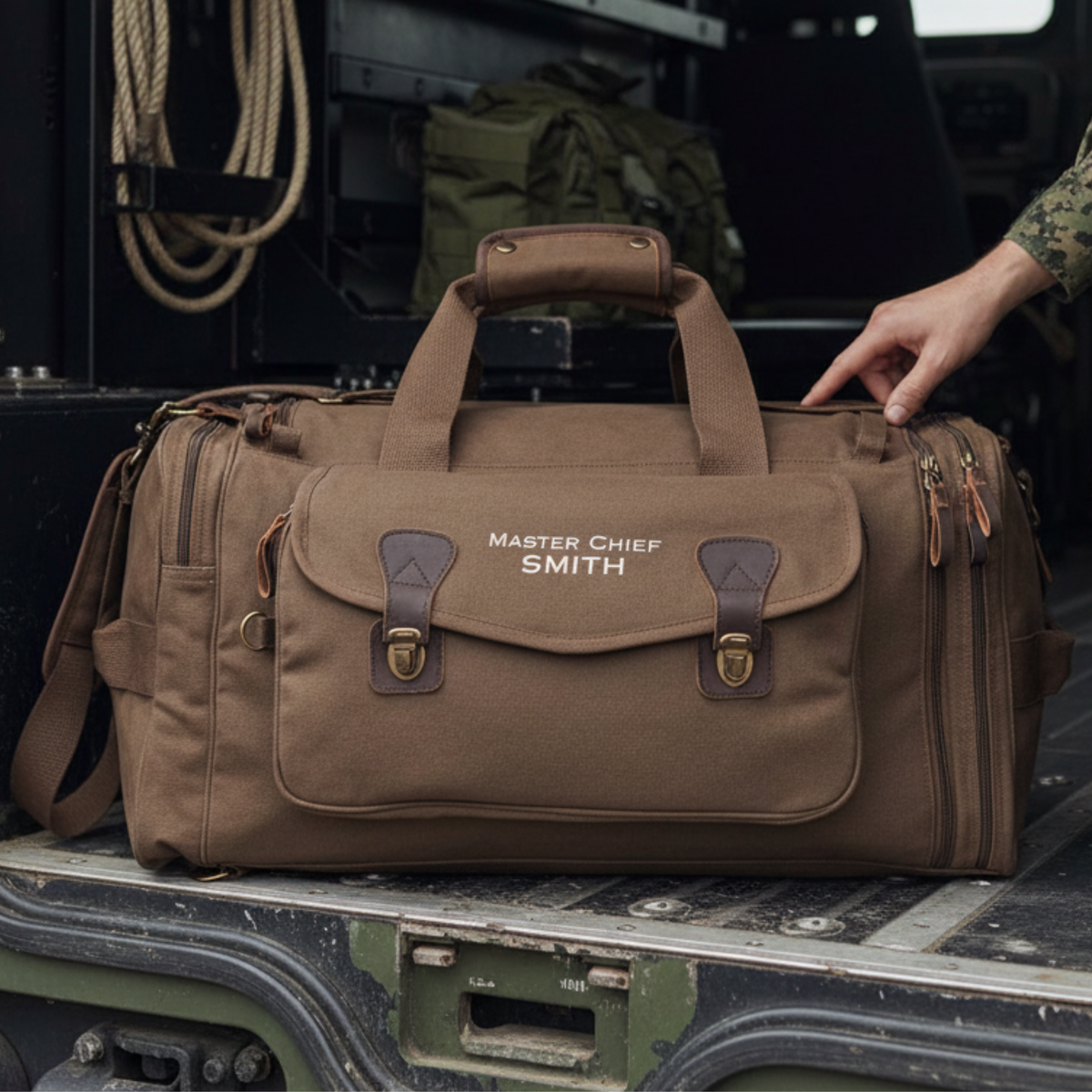 Tan canvas duffel bag personalized with sample text "Master Chief SMITH" and leather trim, displayed on military vehicle with rope and tactical gear in background