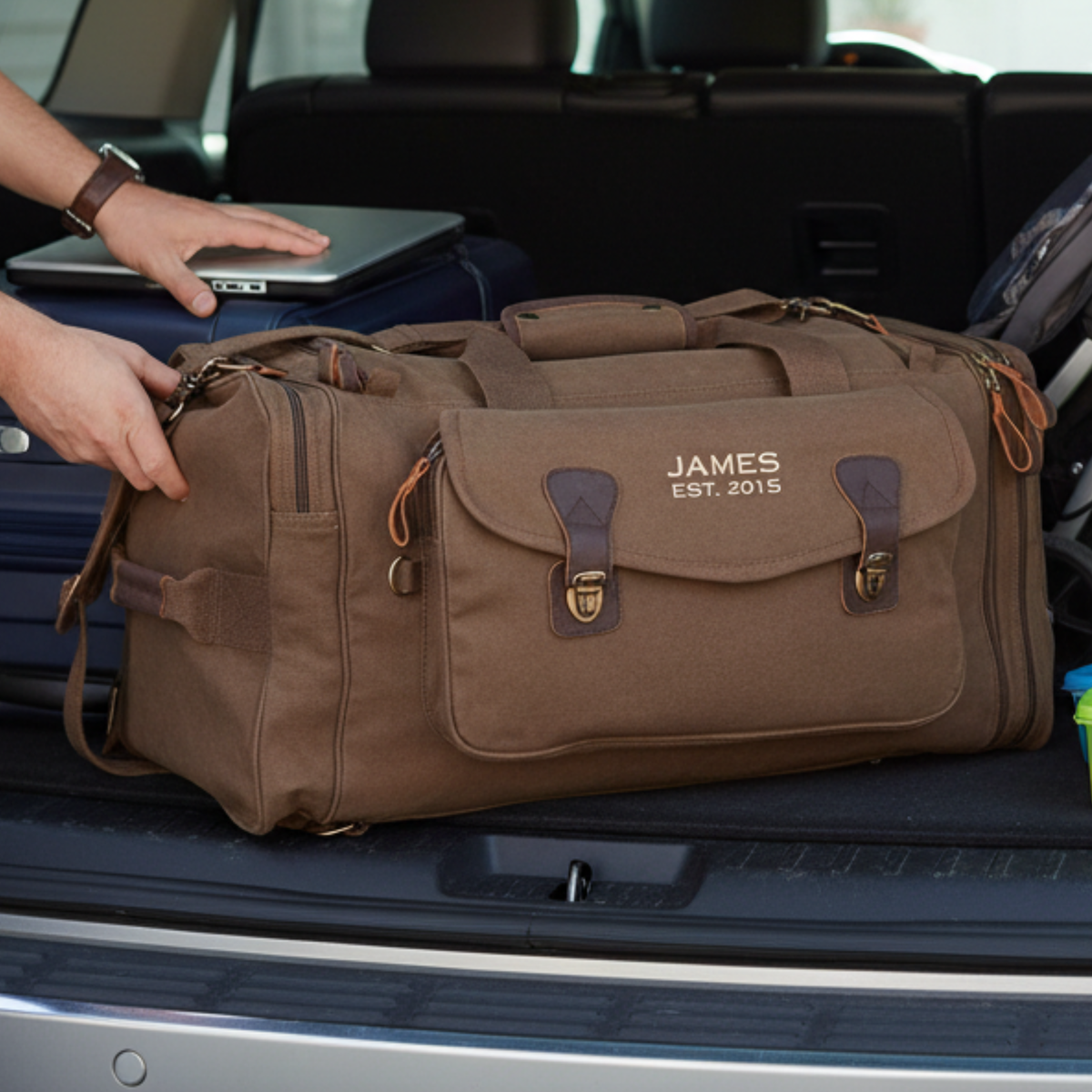 Brown canvas personalized graduation duffle bag embroidered JAMES EST. 2015, loaded into an SUV trunk beside a laptop and suitcase.