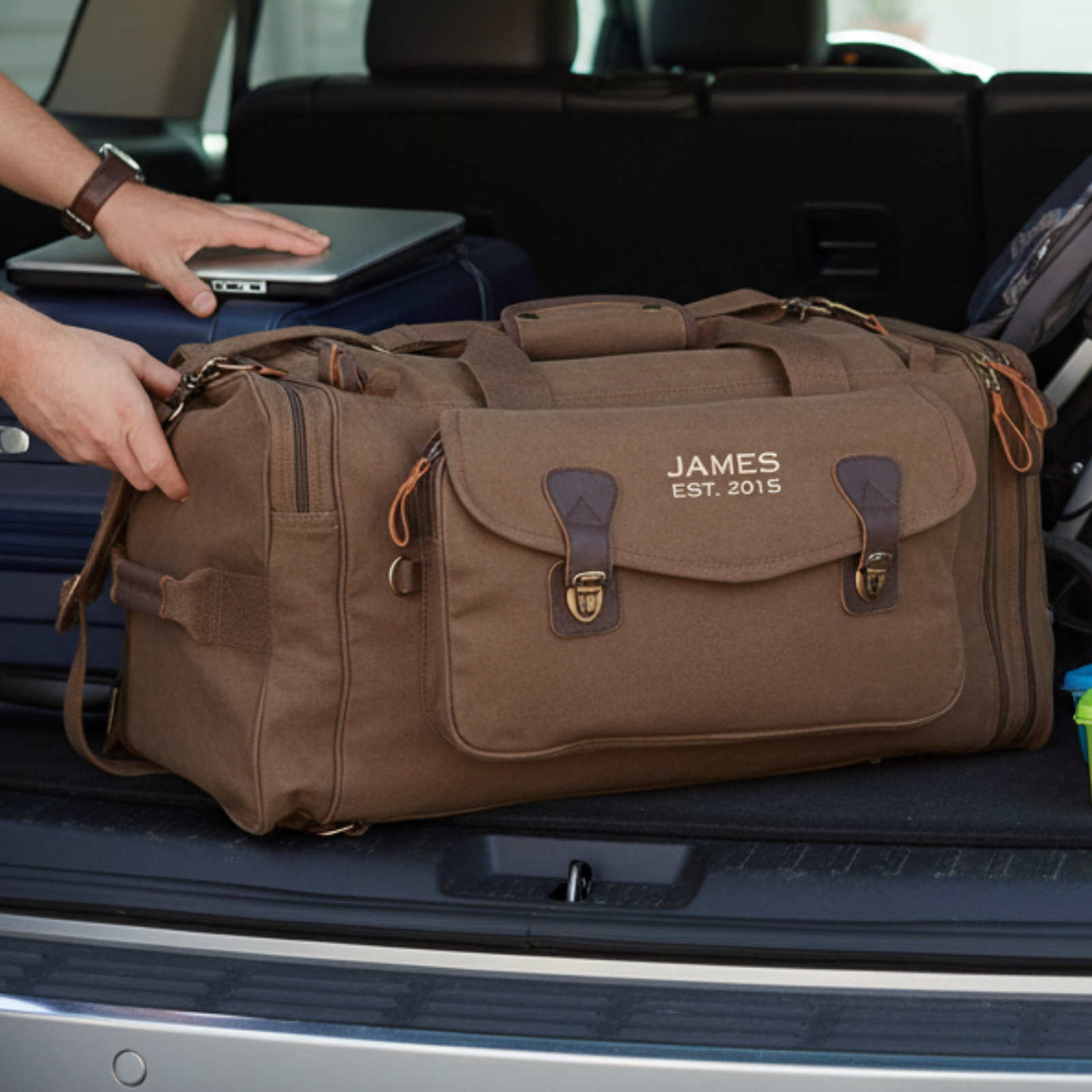 Brown canvas personalized graduation duffle bag embroidered JAMES EST. 2015, loaded into an SUV trunk beside a laptop and suitcase.