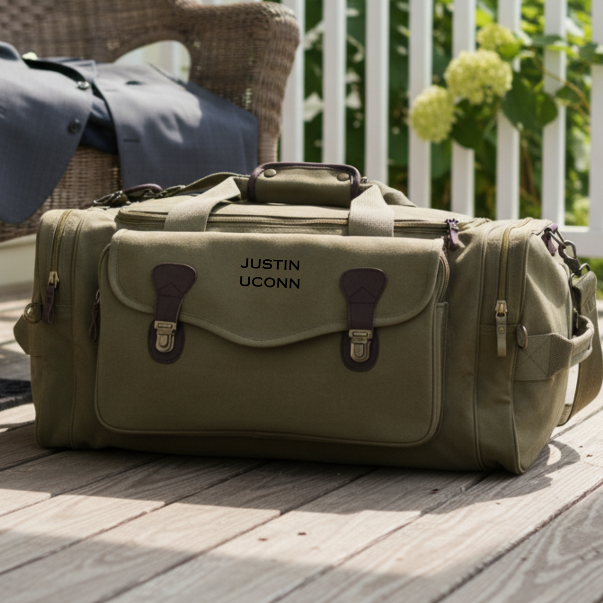Olive green graduation duffle bag embroidered JUSTIN UCONN, on a porch deck beside a wicker chair and a folded dress shirt.