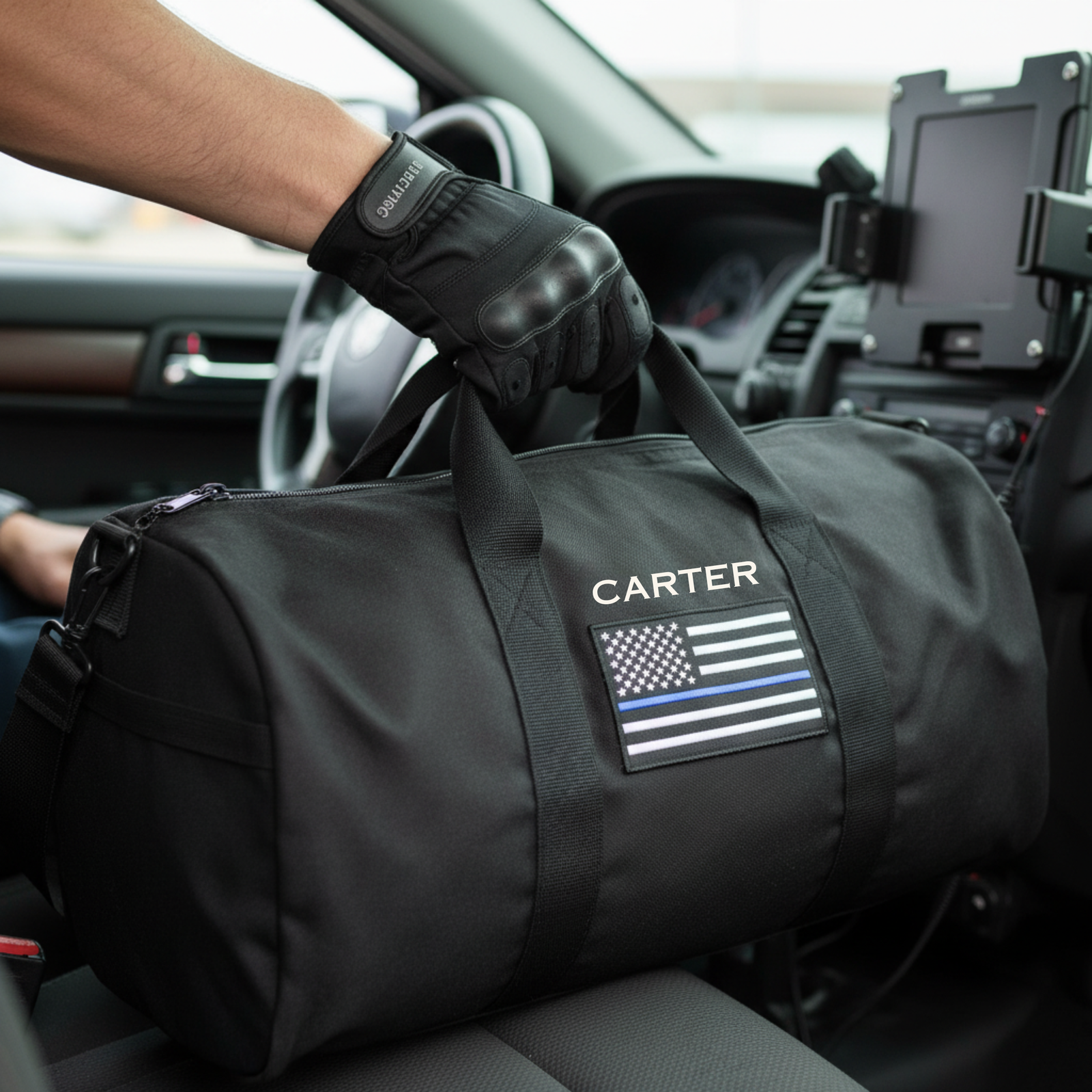 Black canvas duffle bag personalized with sample name "CARTER" and thin blue line flag patch, held in car interior with tactical gloves visible.