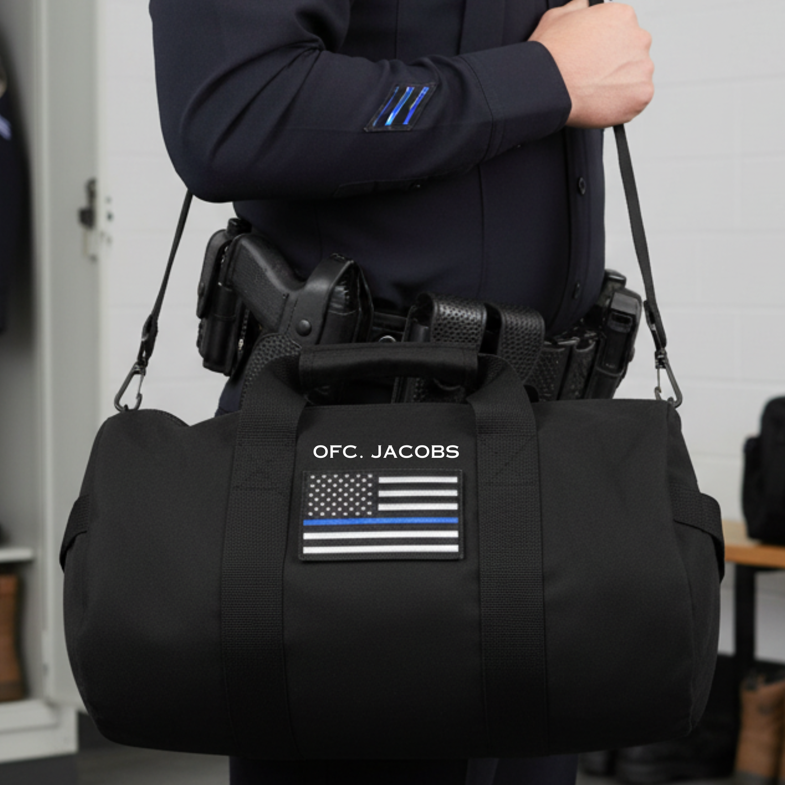 Person wearing navy tactical jacket carrying black canvas duffle bag with personalized name "OFC. JACOBS" and thin blue line flag emblem on front panel