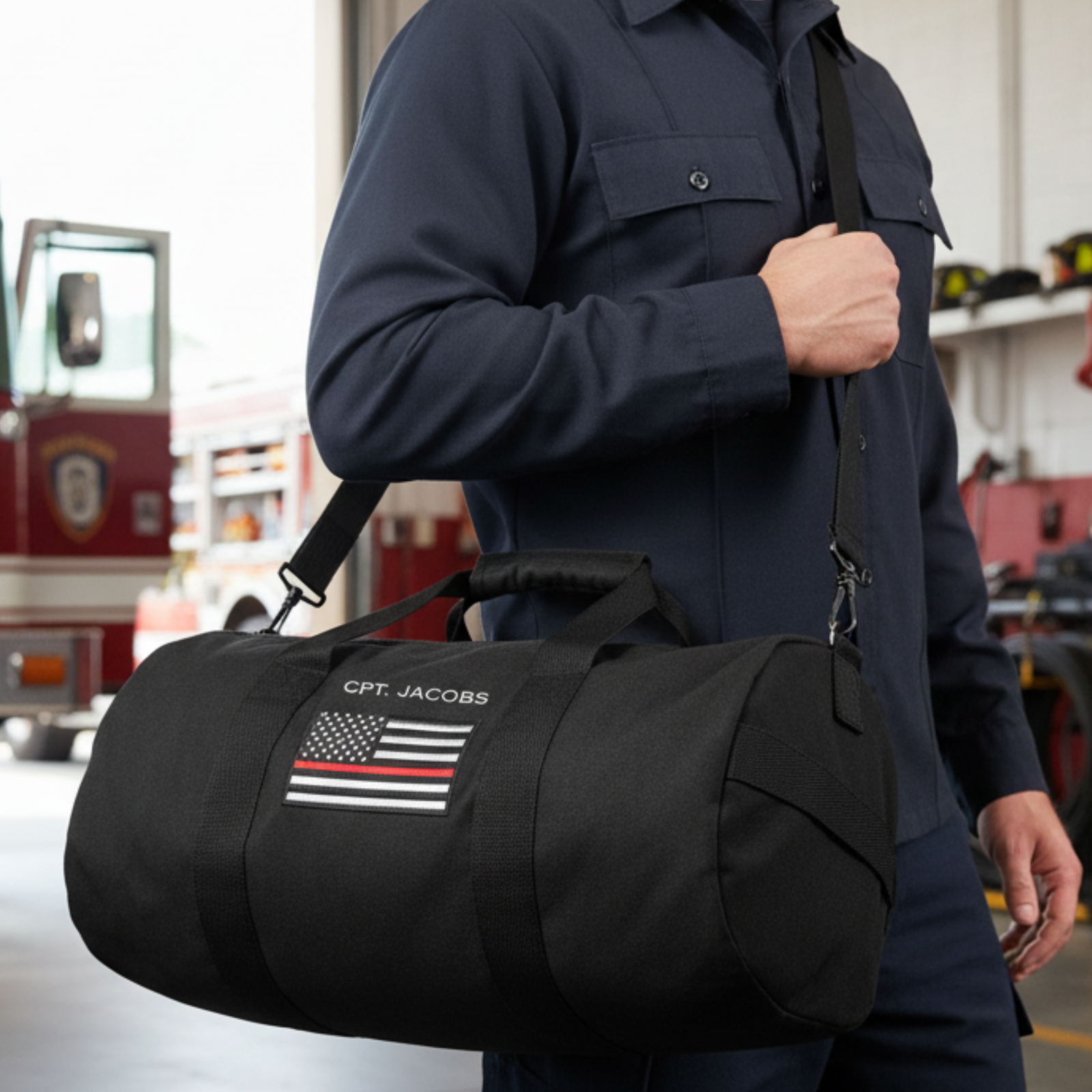 Person wearing navy jacket carrying black canvas duffle bag personalized with sample name "CPT. JACOBS" and thin red line American flag emblem, shown in fire station setting