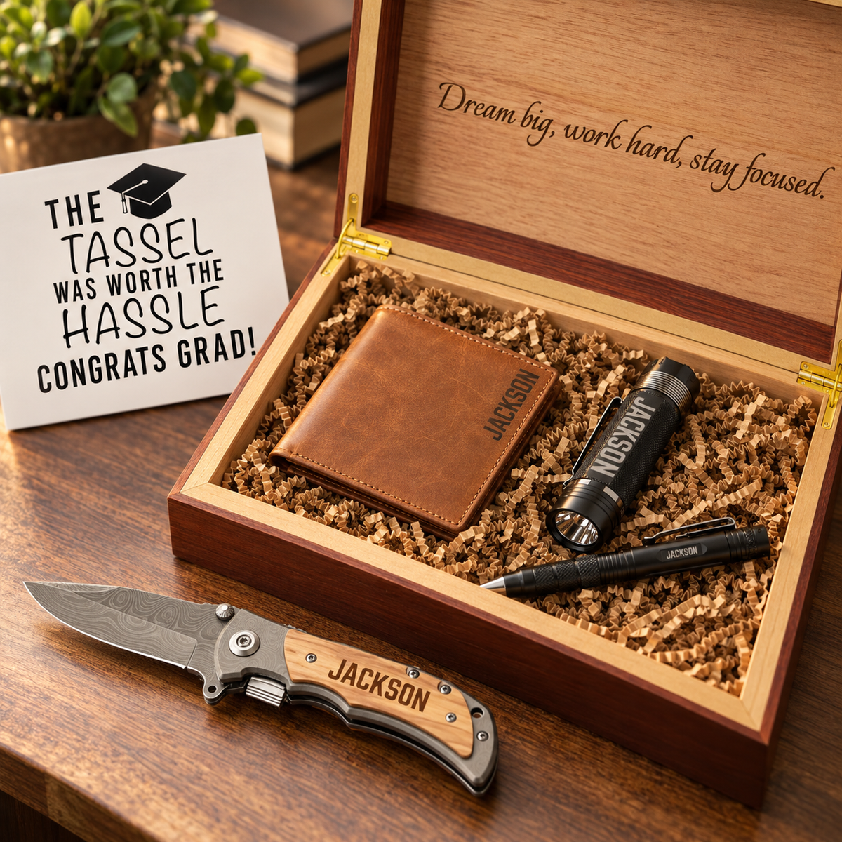 Open wooden box with engraved "Dream big, work hard, stay focused" on lid, containing personalized pocket knife with "JACKSON" engraving, tan leather wallet, black tactical pen, and black flashlight nested in kraft paper, graduation greeting card displayed