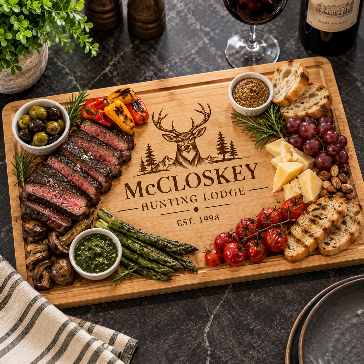 A personalized wooden charcuterie board engraved with 'McCLOSKEY HUNTING LODGE EST. 1998' and a deer silhouette, styled with sliced steak, bread, cheese, grapes, olives, asparagus, mushrooms, and sauces on a dark stone tabletop.