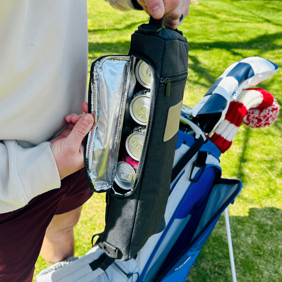 Black neoprene golf cooler bag held open to display four beverage compartments with insulated lining, shown in outdoor setting with golf clubs nearby
