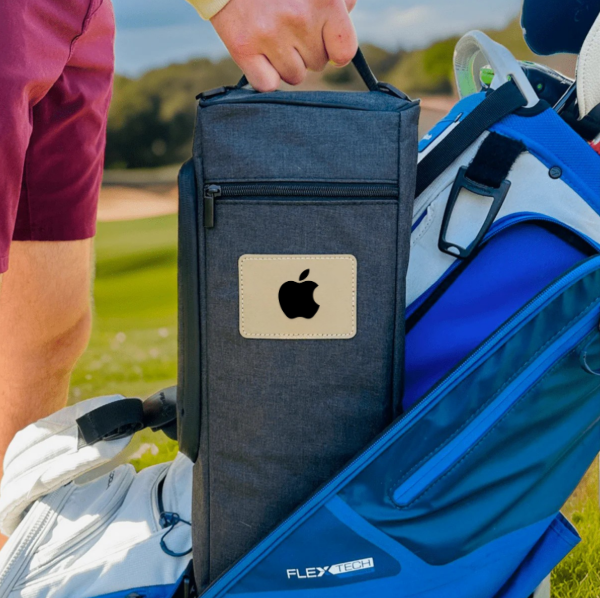 Navy insulated golf cooler bag with cream patch logo being placed in golf bag side pocket, shown on course with golfer wearing pink shirt and blue golf bag visible