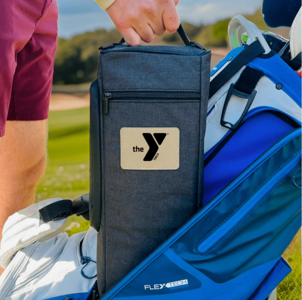 Navy insulated cooler bag with YMCA logo patch being placed into golf bag side pocket on course, surrounded by golf accessories and blue golf bag.