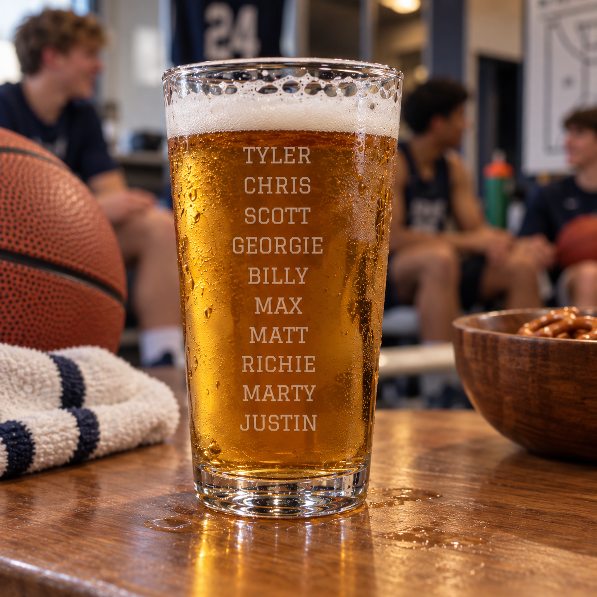 Tall pint glass filled with beer, engraved with sample names Tyler, Chris, Scott, Georgie, Billy, Max, Matt, Richie, Marty, Justin on wood table with basketball and snacks
