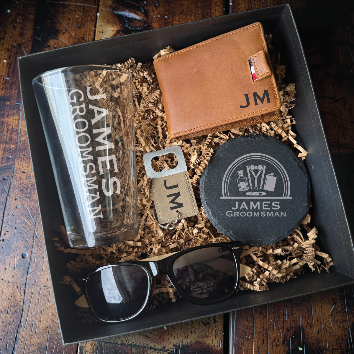 Personalized gift box for him in a black box with crinkle fill, featuring an engraved pint glass, leather wallet with sample initials JM, coaster, sunglasses, and bottle opener.
