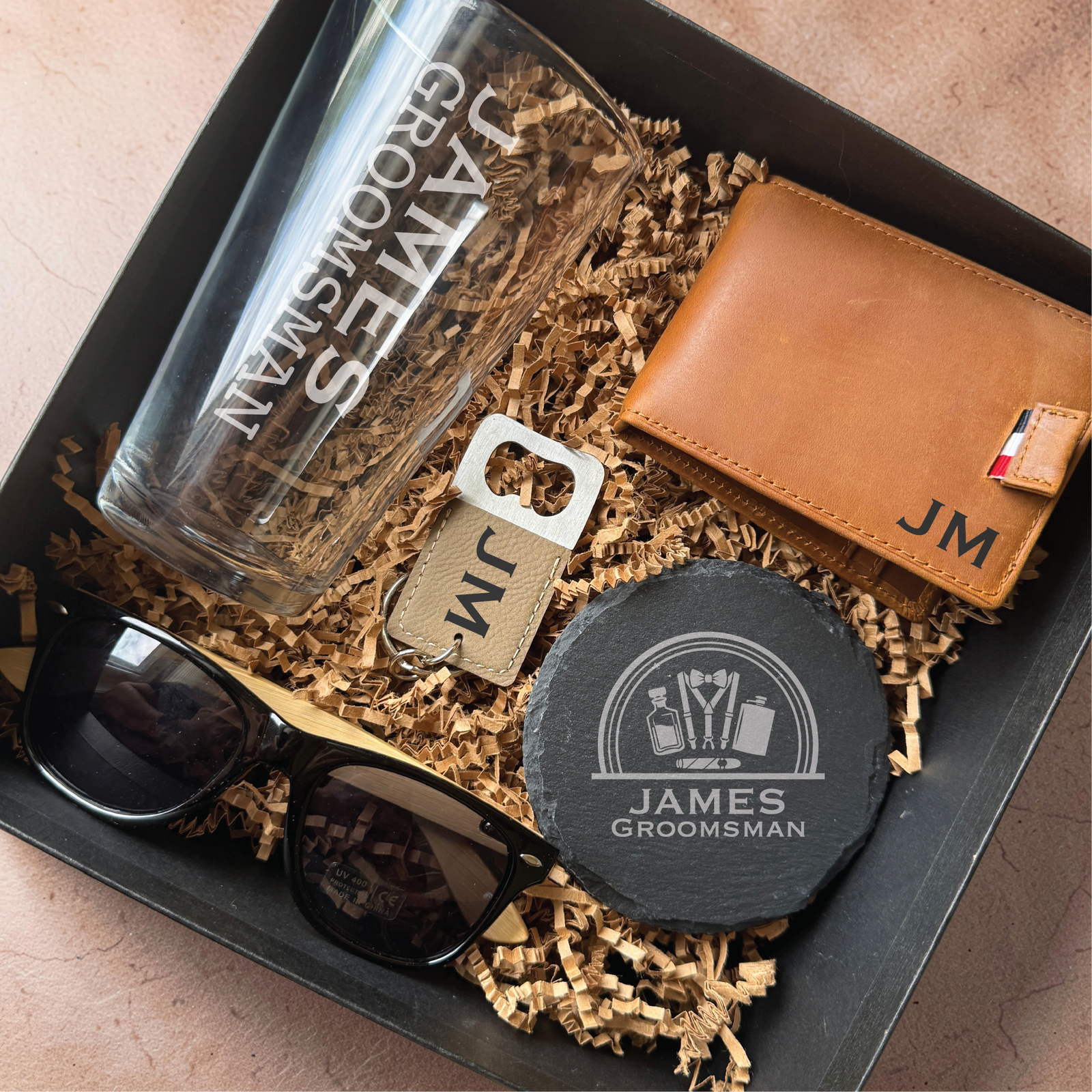 Top view of a personalized gift box for him with gold crinkle fill, engraved pint glass, leather bifold wallet, slate coaster, and bottle opener keychain with sample initials JM.