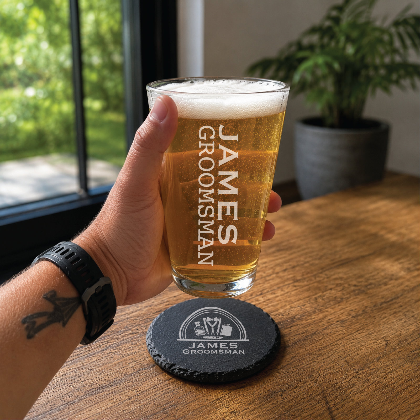 Hand holding an engraved pint glass beside a round slate coaster with sample name James, from a personalized gift box for him.
