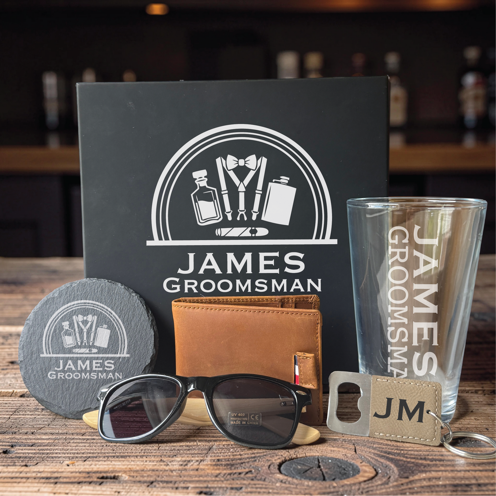 Personalized men's gift box with engraved pint glass, tan leather wallet, slate coaster, polarized sunglasses, and JM bottle opener keychain, sample name James on a bar.