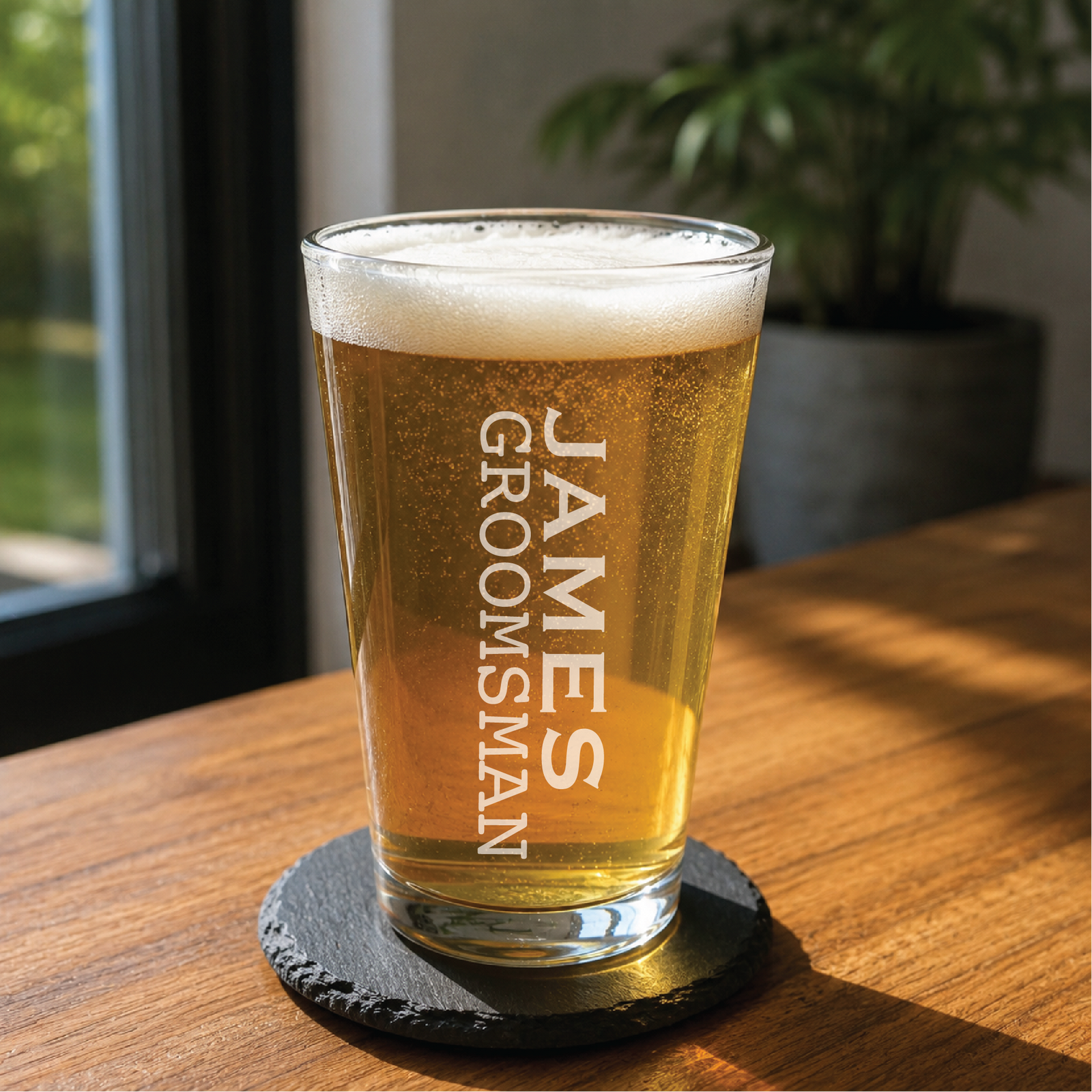 Engraved 16 oz pint glass with sample name James filled with beer, resting on a slate coaster, part of a personalized men's gift set.