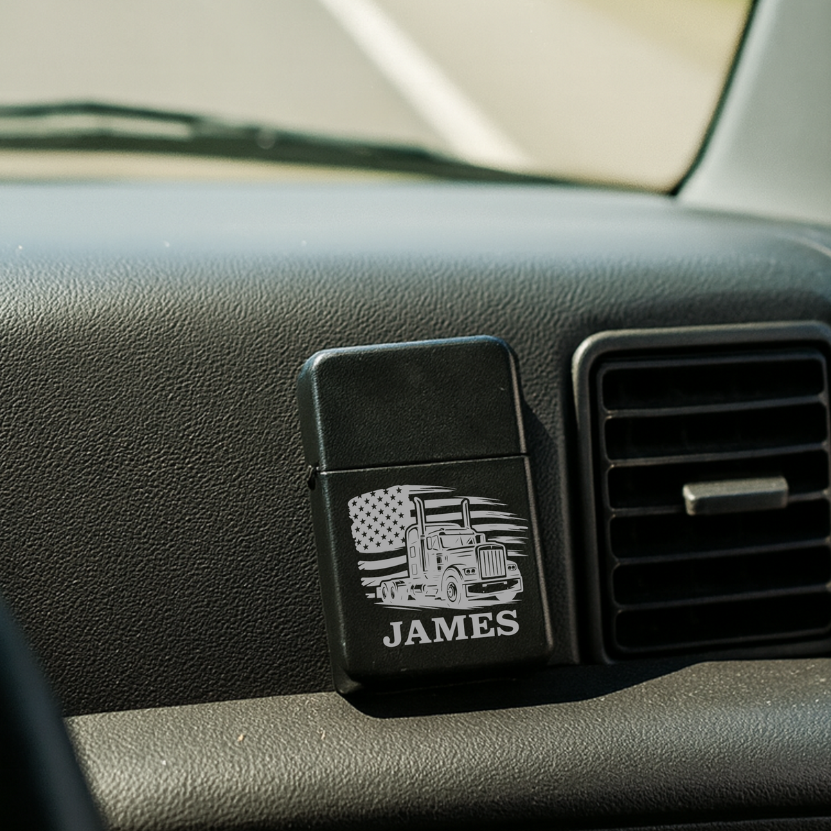 Black flip lighter displayed on car dashboard with personalized engraving showing sample name "JAMES," semi-truck graphic, and American flag design