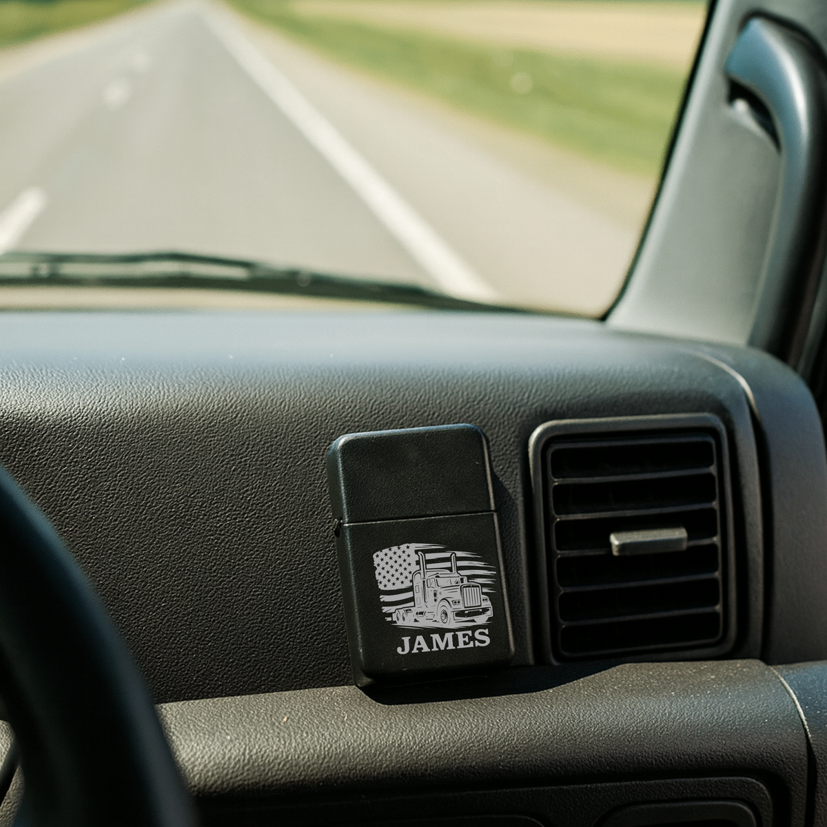 Personalized flip lighter mounted on vehicle dashboard, laser-engraved with sample name "JAMES" and semi-truck with American flag design, shown in car interior setting.