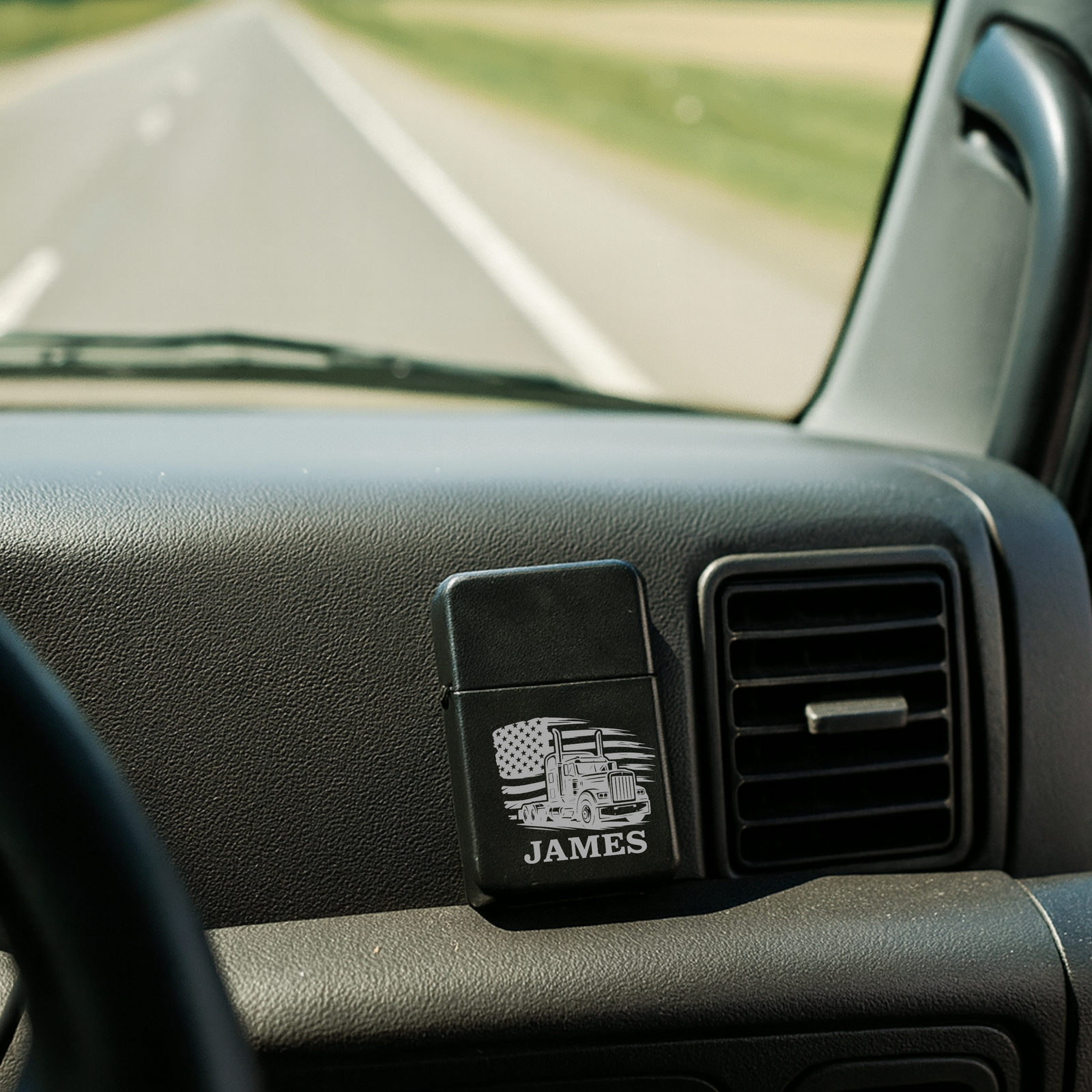 Personalized flip lighter mounted on vehicle dashboard, laser-engraved with sample name "JAMES" and semi-truck with American flag design, shown in car interior setting.