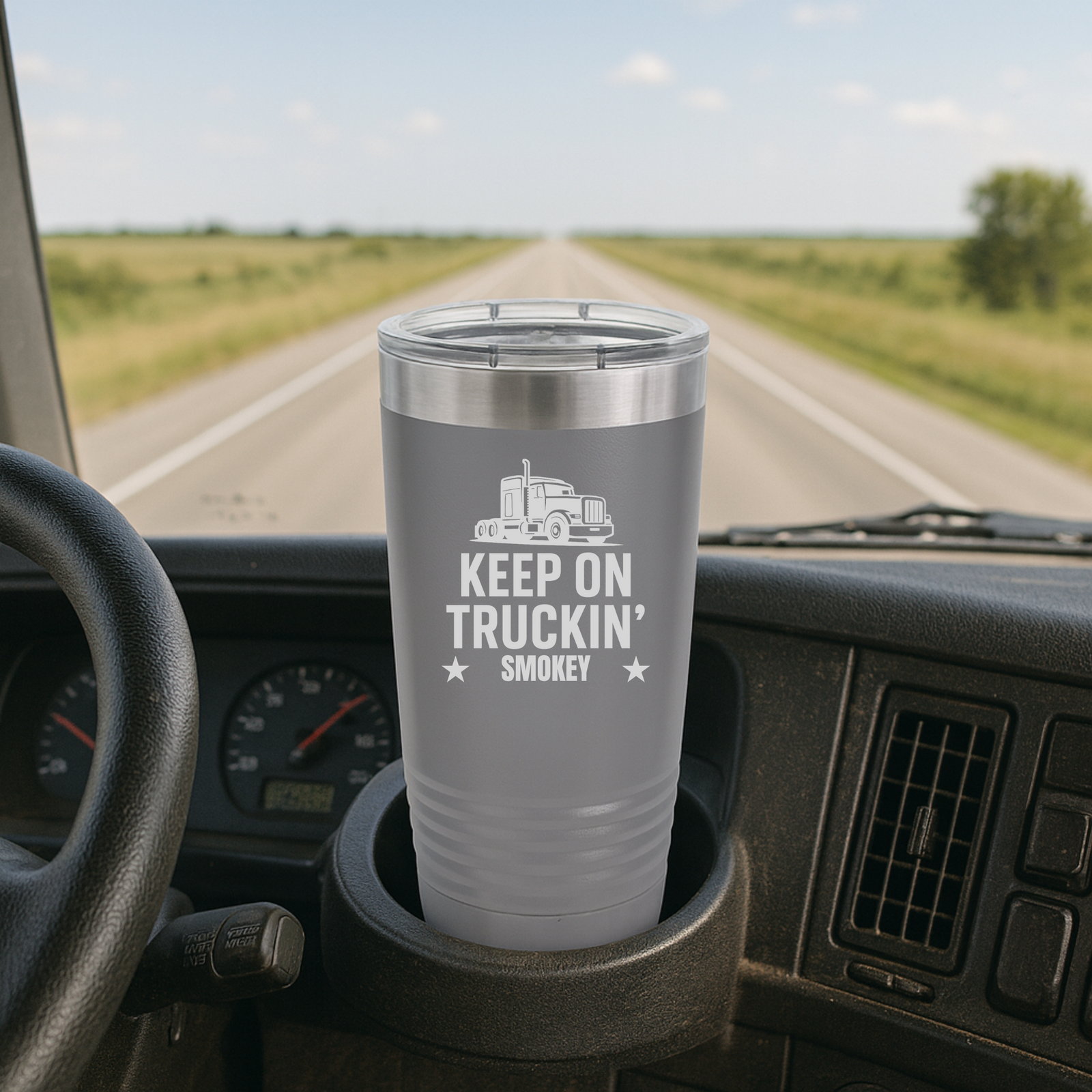 Gray insulated tumbler with "KEEP ON TRUCKIN' SMOKEY" text and truck graphic, positioned in truck cab cup holder with highway stretching ahead