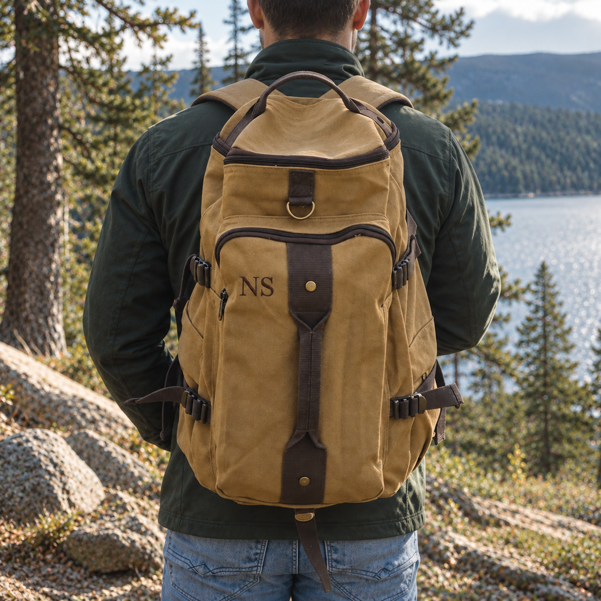 Man by a mountain lake wearing a brown canvas convertible backpack with leather trim, embroidered with sample initials NS on the front pocket