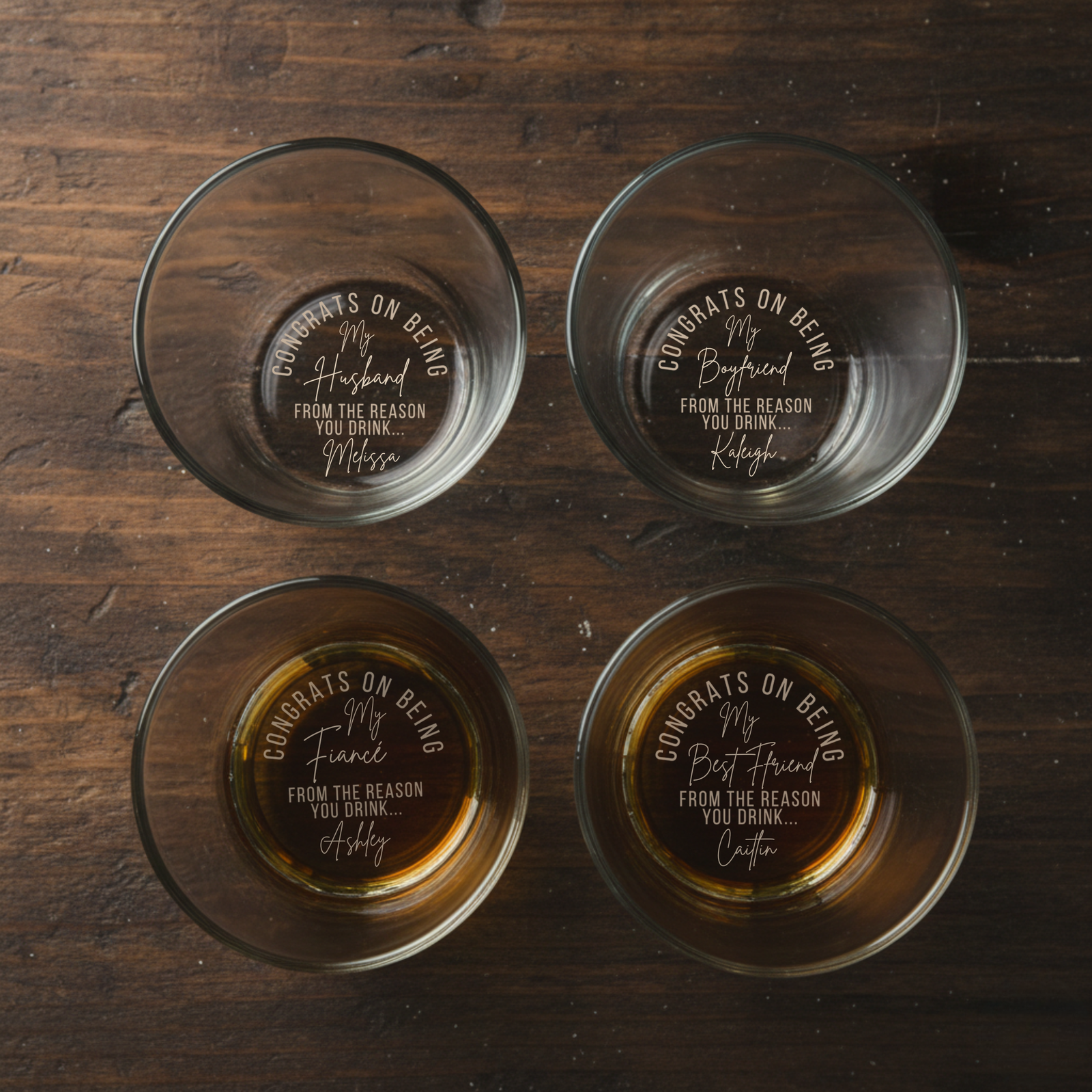 Four whiskey glasses arranged overhead on wood surface, each laser-engraved with "Congrats on Being" and sample personalized names with "From the Reason You Drink" text