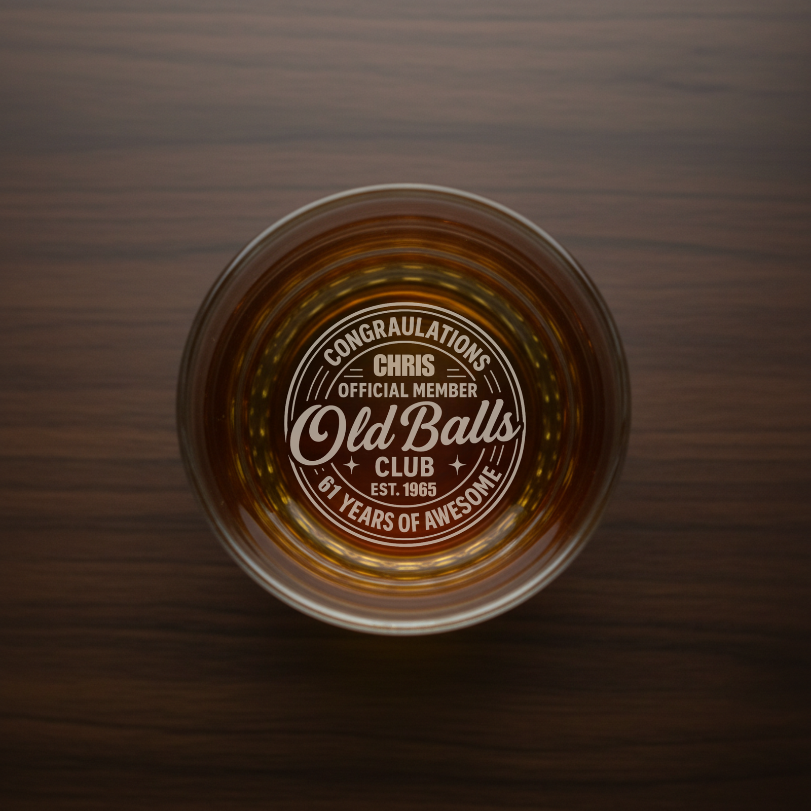 Overhead view of whiskey glass filled with amber liquid, showing laser-engraved "Old Balls Club" design and sample name "Chris" on bottom