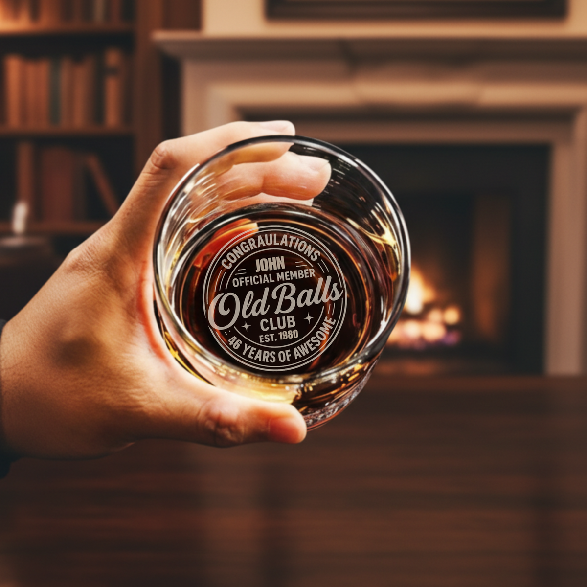 Hand holding whiskey glass filled with amber liquid, laser-engraved with sample personalization reading "Congratulations John Official Member Old Balls Club Est. 1980 46 Years of Awesome," fireplace blurred in background