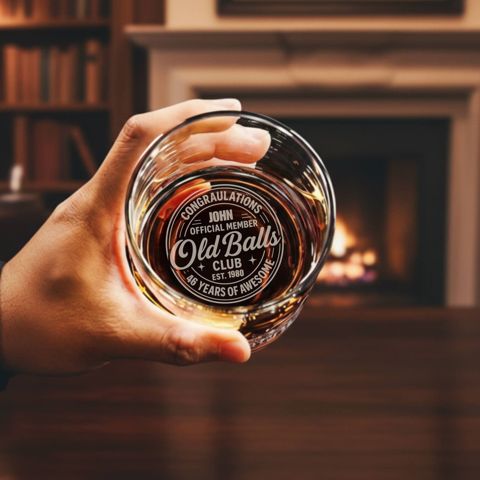 Hand holding whiskey glass filled with amber liquid, laser-engraved with sample personalization reading "Congratulations John Official Member Old Balls Club Est. 1980 46 Years of Awesome," fireplace blurred in background
