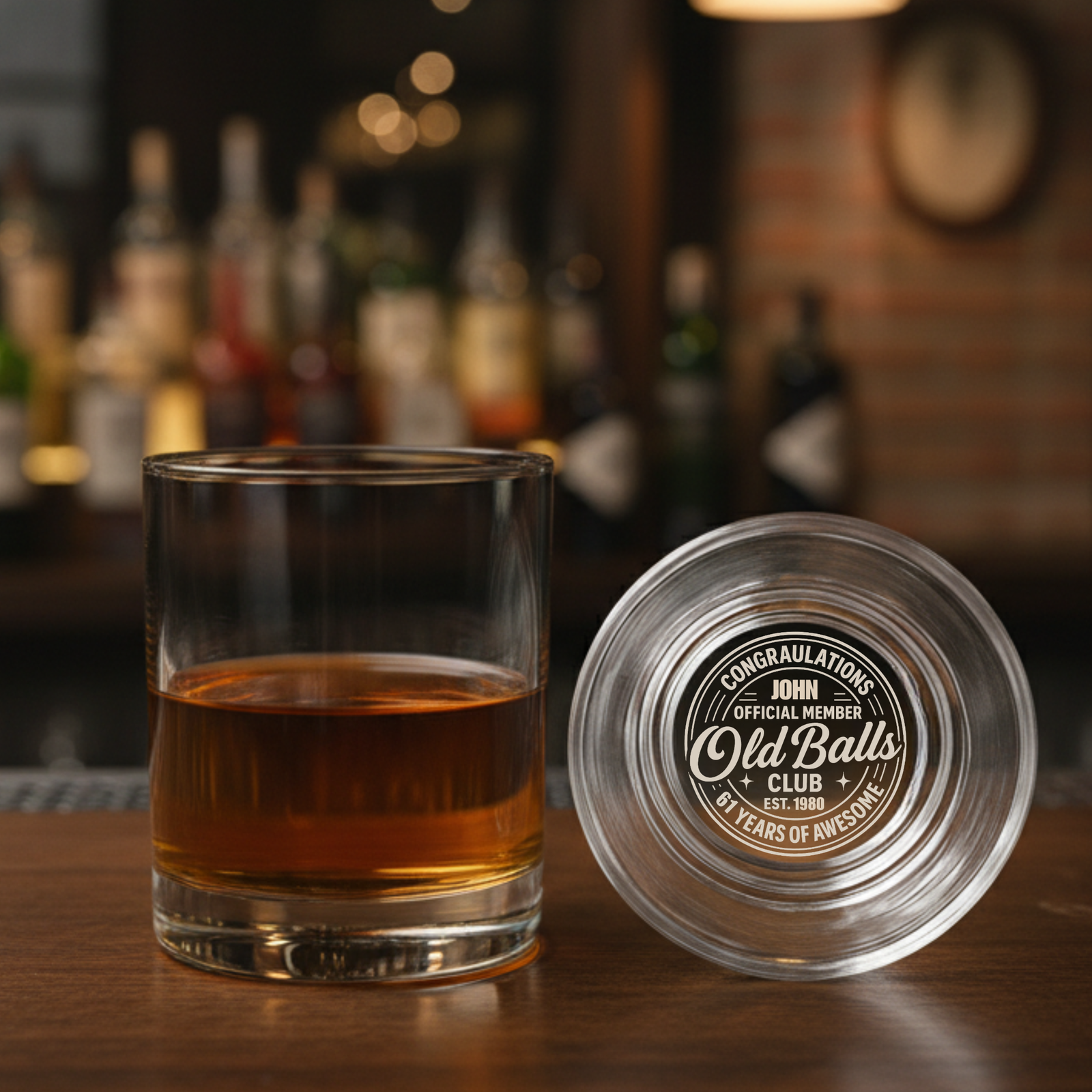 Whiskey glass filled with amber liquid next to matching coaster, both laser-engraved with sample "Old Balls Club" design and "Congratulations John" text on wooden bar surface